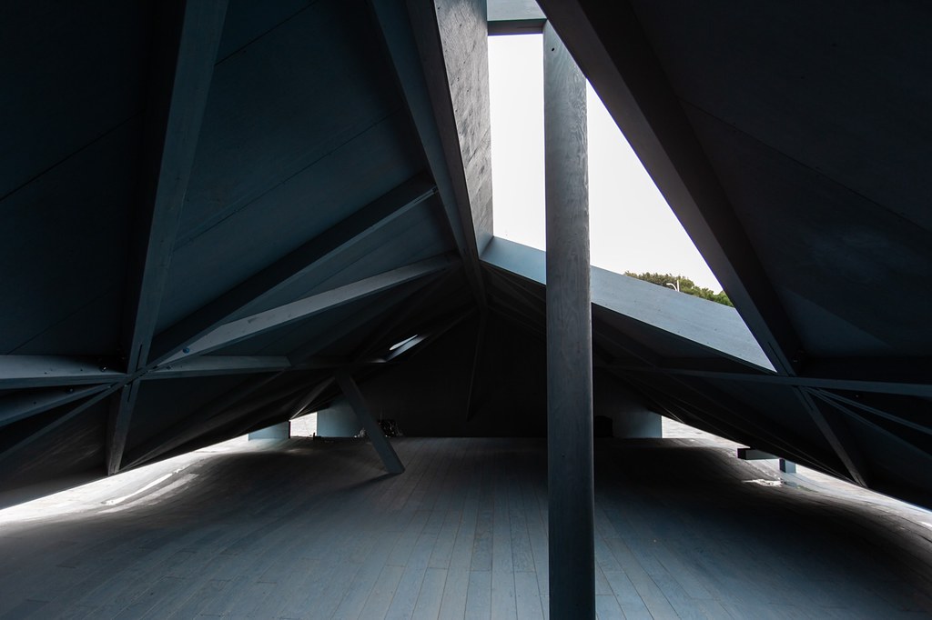 《藍屋 Blue House》團隊刻意在不規則的輪廓間刻畫出建築物的穿透性。如屋簷間留下的隙縫、與斜屋頂上的幾何開孔等透過木作細節的抽象表徵，再次模糊、霧化真實與虛構的邊界。而詩化意象與觀眾身體間交互營造的遊走經驗，也延展出空間的複調敘事，終而形成一種兼具普遍性及多元性的建築形態。