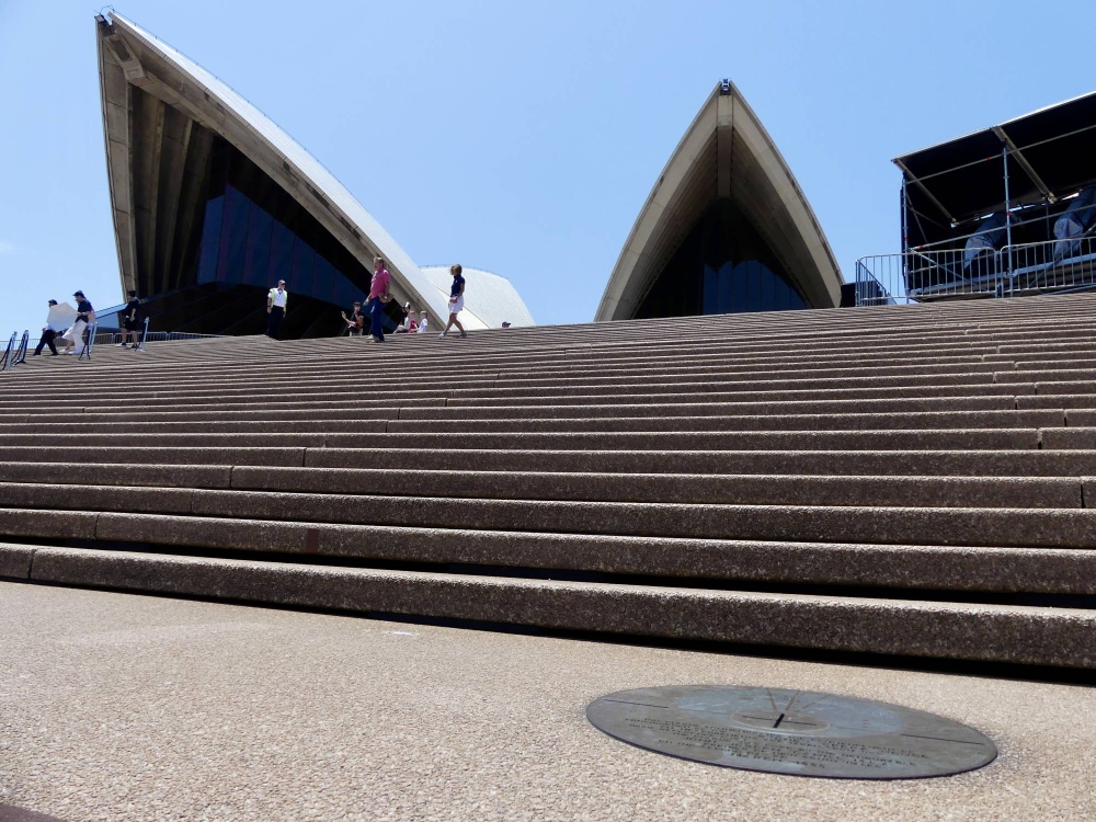 A bronze plaque with a diameter of 61 centimeters, which marks the point where the axes of the two halls intersect, can be found at the bottom of the steps leading to the two halls of the Sydney Opera House. 在通往雪梨歌劇院兩個大廳的台階底部，有一塊直徑為 61 釐米的銅牌，標誌著兩個大廳軸線的交點。| TOMOKO OTAKE