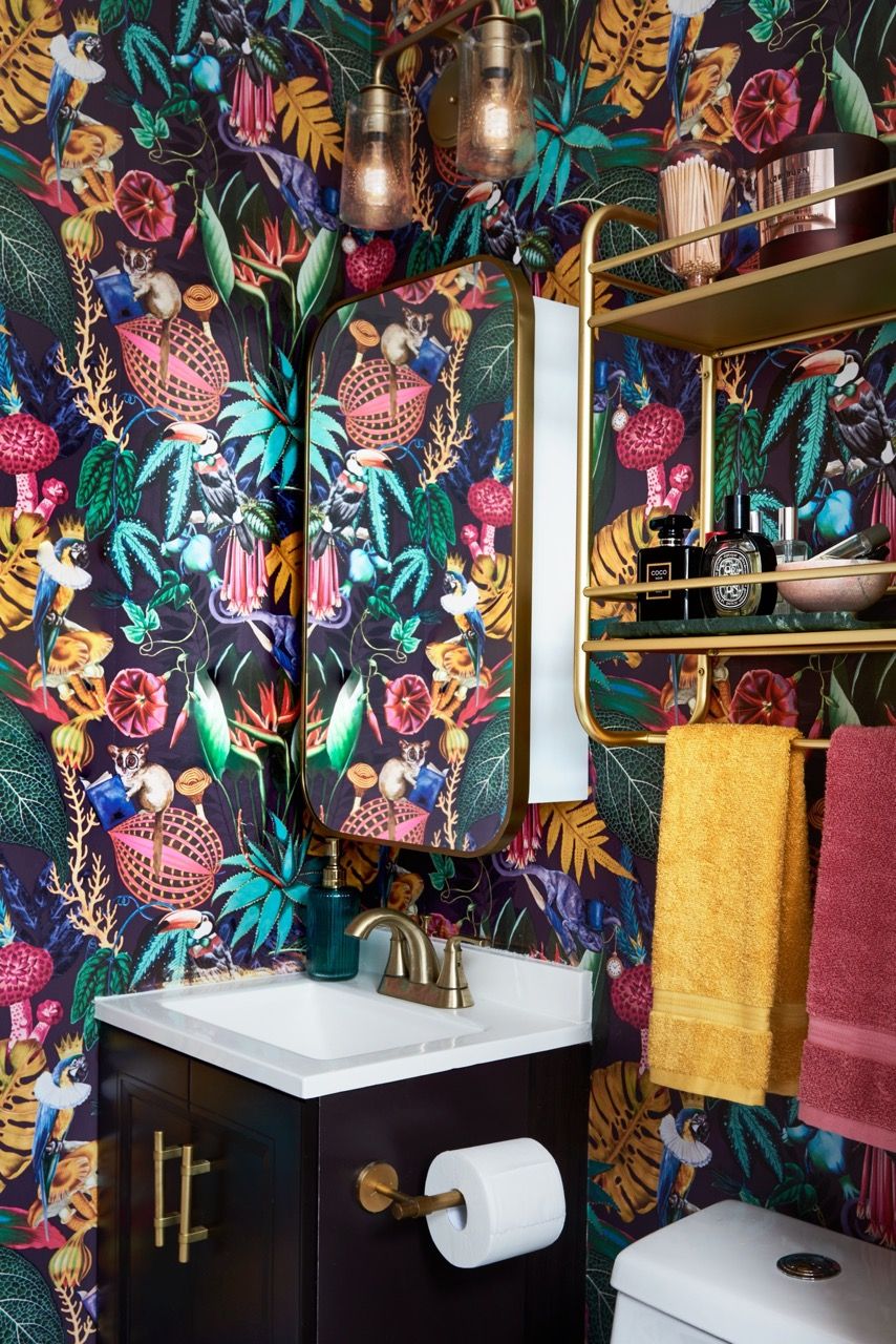 a bathroom with a wall of art©Marc Mauldin Photography 在Blessed Little Bungalow設計的這間洗手間中，壁紙上重複的寶石色調與梳妝台、鏡子和馬桶上方架子上的金色飾面完美搭配