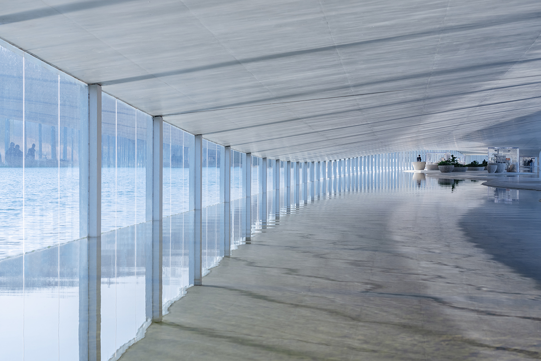 在水美術館 石上純也 Junya Ishigami 在「在水美術館」的長廊下，光影與反射編織出一幅瞬息萬變的風景。透明的牆面將外界的景致一一攬入，每個角度都呈現不同的視覺詩篇。游客漫步於此，如同行走在水的另一端，與遠處的城市景觀相望，空間感被極致放大。光線穿透屋頂的纖薄，於灰色的混凝土上投下柔和的圖案，每一步都踏在光的節奏上。