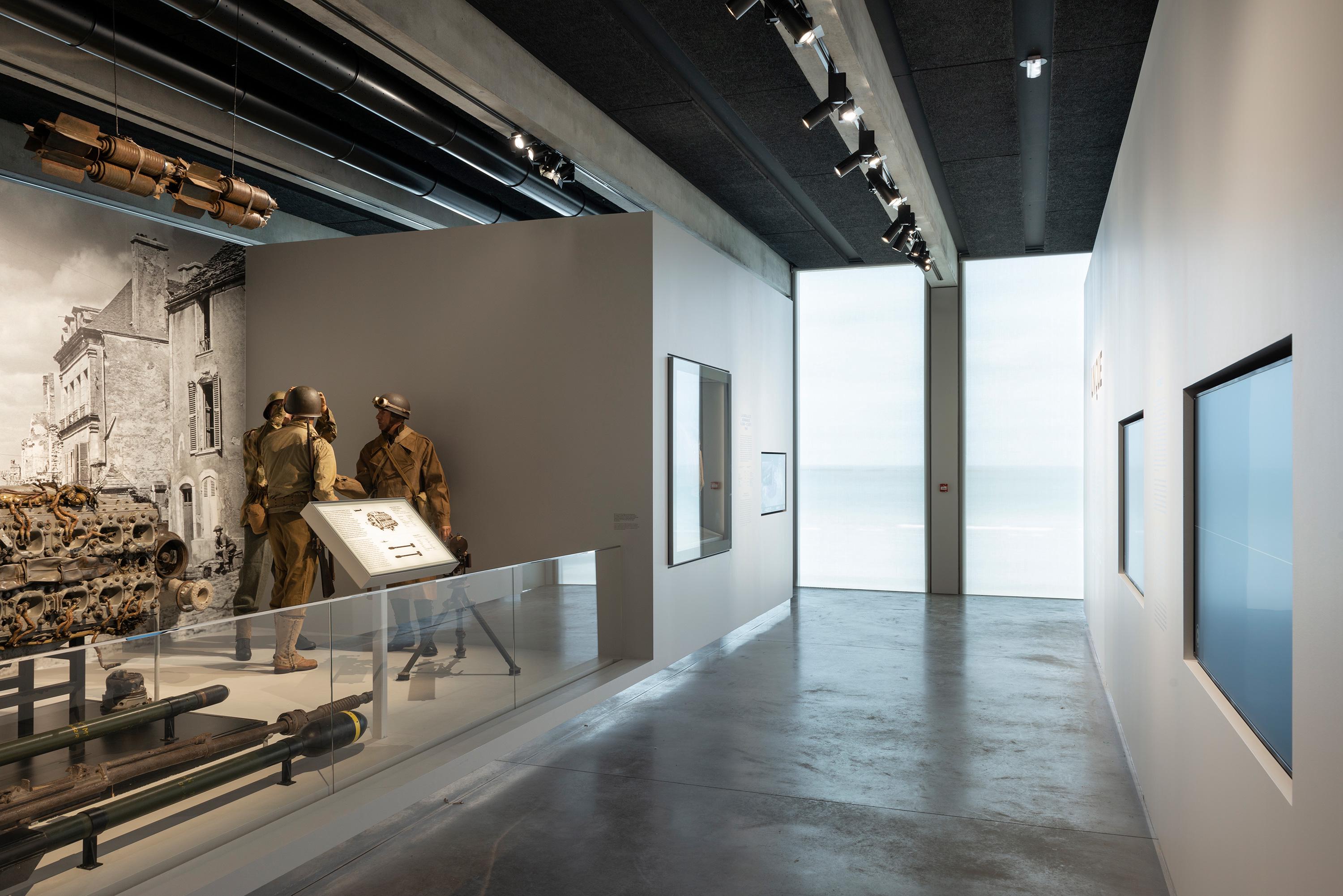 PROJECTILES, Antoine Cardi · D-Day Museum 博物館內部延續了外部的預製混凝土結構，12米長的橫梁以2米的間隔排列，從中央的「裂縫」延伸至南北立面。這種結構設計不僅提供了穩固的支撐，更創造出開闊、流動的空間感，讓參觀者在欣賞展品的同時，也能感受到建築本身的力量與美感