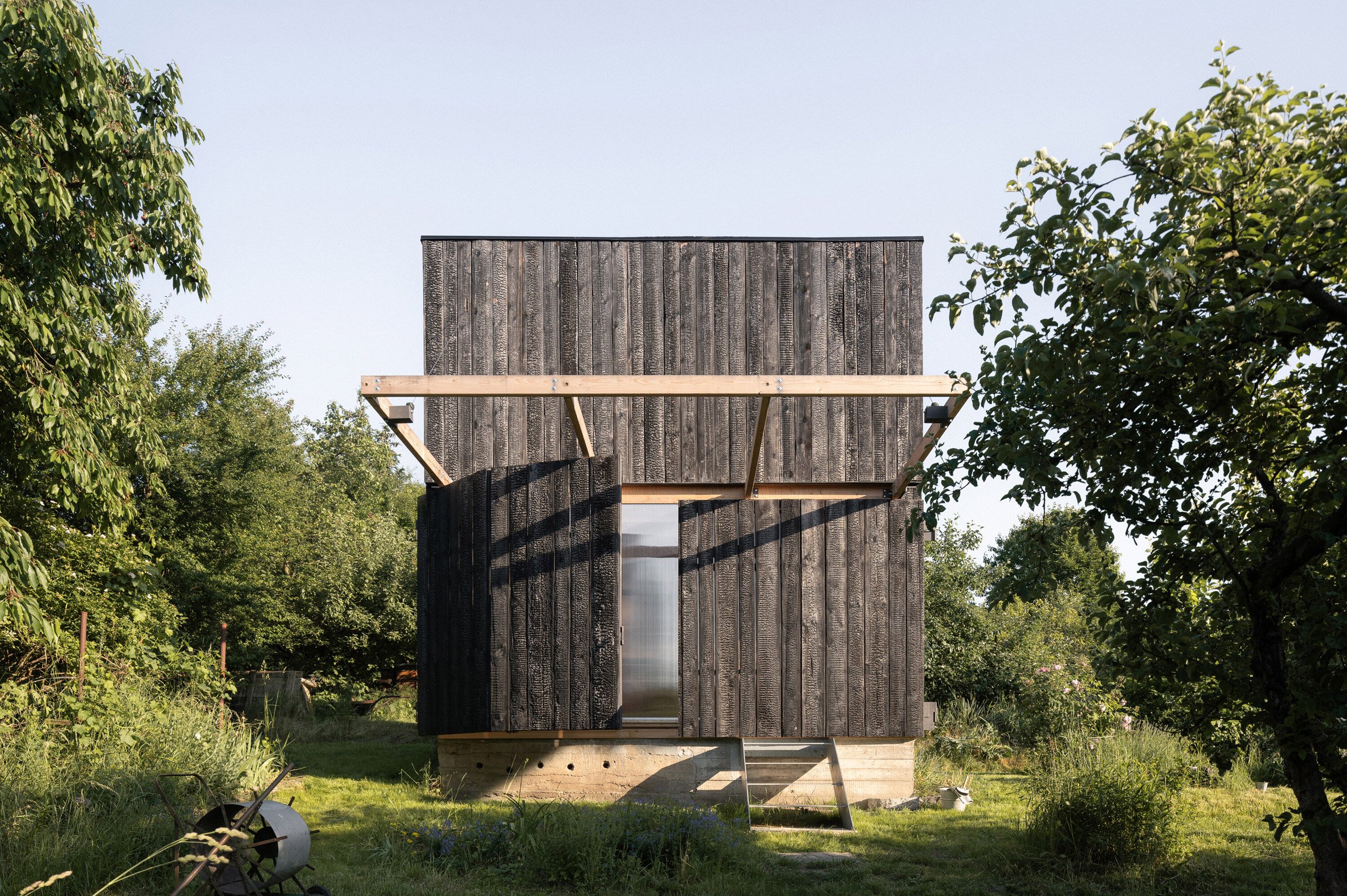 Byró Architekti 設計團隊希望這座小木屋能與周圍的花園融為一體，因此特別設計了一面可開闔的PC板牆。這面牆隱藏在木百葉窗後方，開啟時能將室內空間延伸至戶外，同時形成一個遮蔽的戶外空間，讓使用者在雨天也能享受花園的氛圍