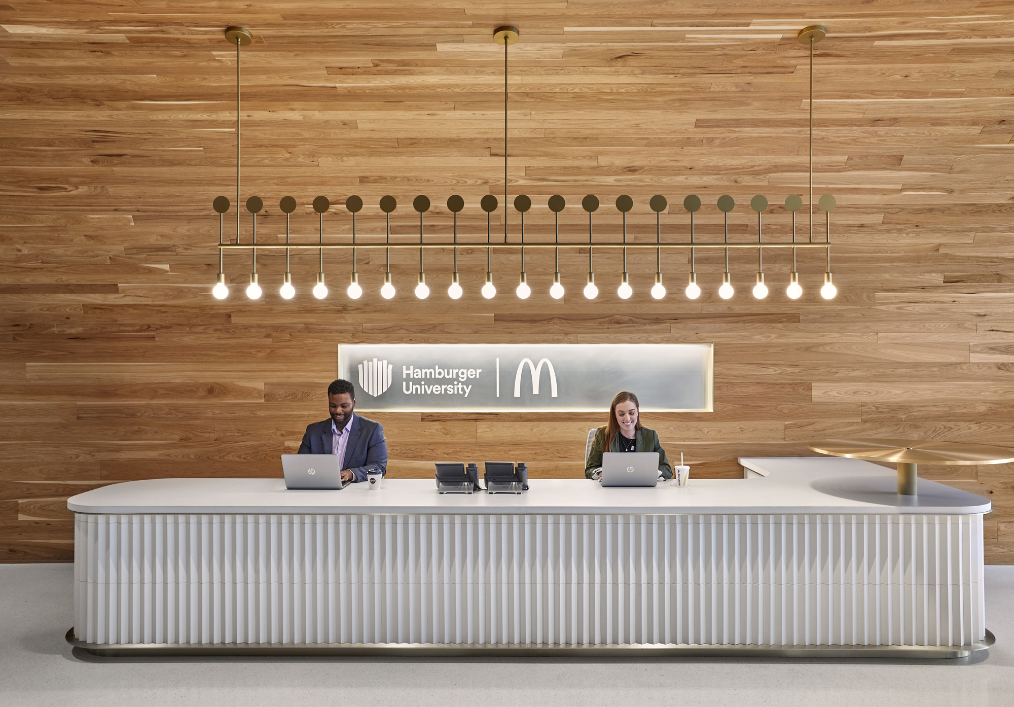 芝加哥麥當勞企業總部 Hamburger University 接待區採用溫暖木材覆蓋背景牆，配合發光品牌標誌突顯專業形象。白色接待桌以流線立體設計呈現，搭配金屬細節提升質感。上方懸掛對稱排列的金屬燈具，營造簡潔而明亮的氛圍，強化空間的迎賓功能