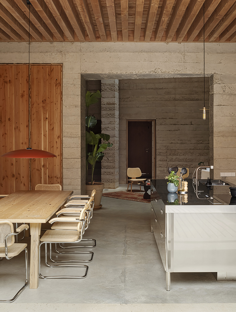 餐廳與廚房空間搭配裸露的天花，呈現出樸實而溫潤的對比。實木餐桌與經典的 Cesca 藤編椅（由 Marcel Breuer 設計）帶來現代主義的細膩觸感，金屬管狀結構增添輕盈感。廚房中島採用不鏽鋼材質，質感俐落且實用，與自然元素相映成趣。橙色吊燈為空間增添暖色點綴