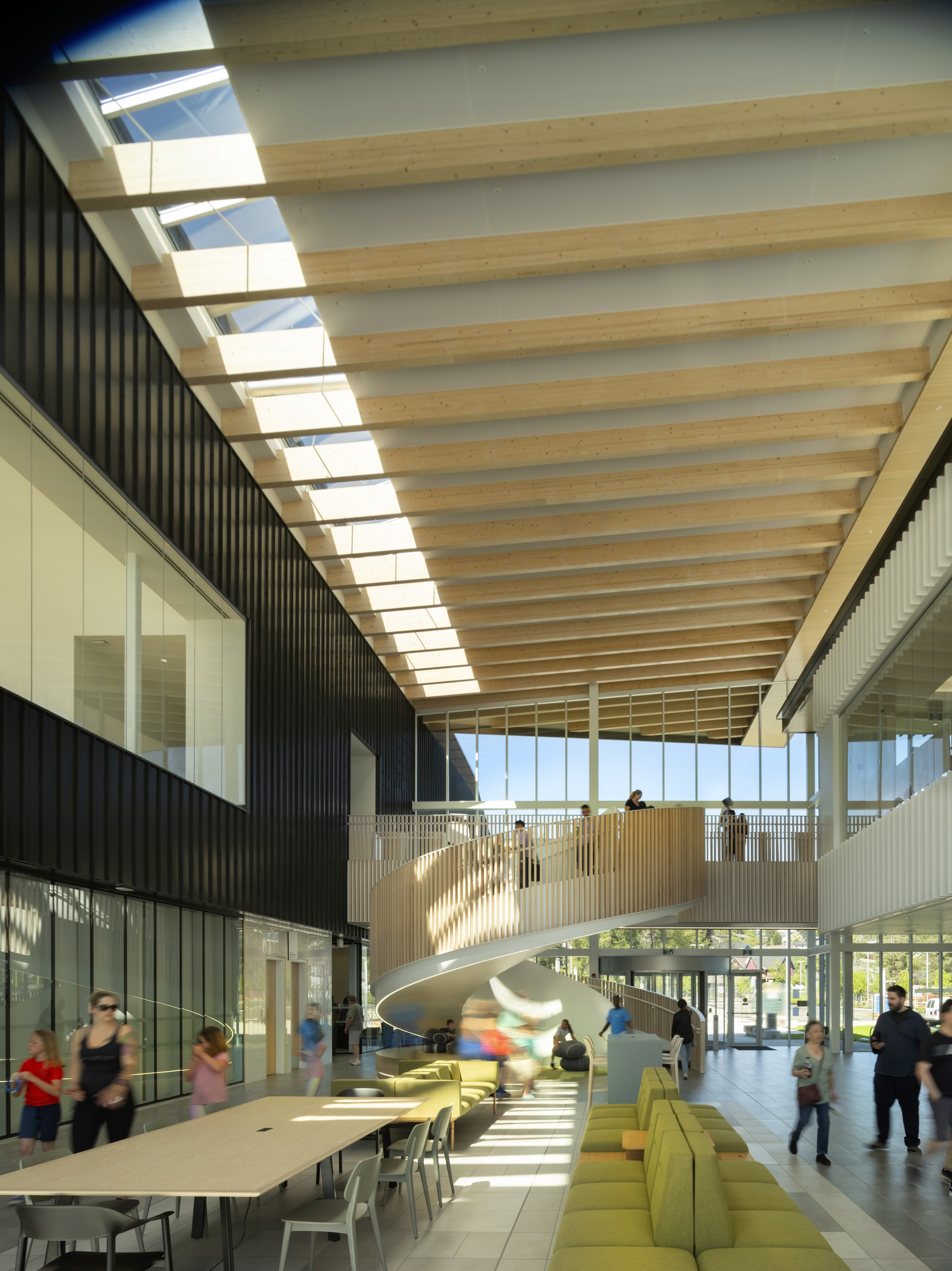 təməsew̓txʷ水上運動中心暨社區中心內部以自然採光與木頭材質強調舒適氛圍，螺旋樓梯成為視覺焦點，木欄杆與白色結構對比鮮明。天花板採用膠合木樑結構，讓光影隨時間變化展現動態美感；Muuto品牌家具，簡約設計與綠色布料座椅增添空間活力。開放式中庭設計促進人流互動，為使用者提供多功能與歡迎的公共空間