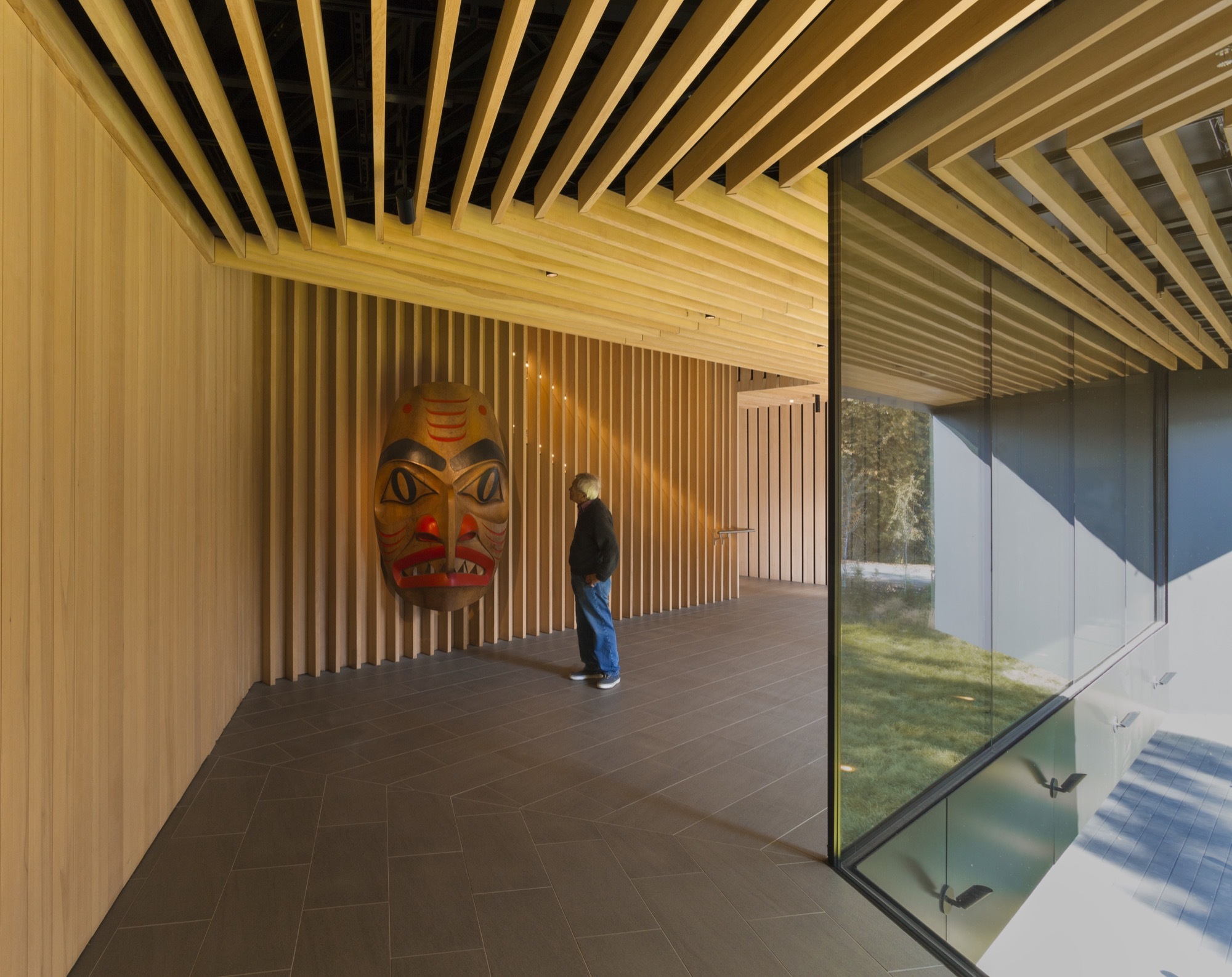 Patkau Architects 加拿大奧登美術館 Audain Art Museum 室內展區採用天然木材作為牆面和天花板的主要材質，細緻排列的木質格柵延伸空間感，營造溫暖且靜謐的氛圍。展示牆上懸掛一件原住民藝術品——大型傳統面具，展現加拿大西岸原住民族文化的獨特魅力。外牆的大片玻璃提供自然光線，將室內與森林景觀連結，使藝術品的觀賞經驗更具空間延續性與沉浸感