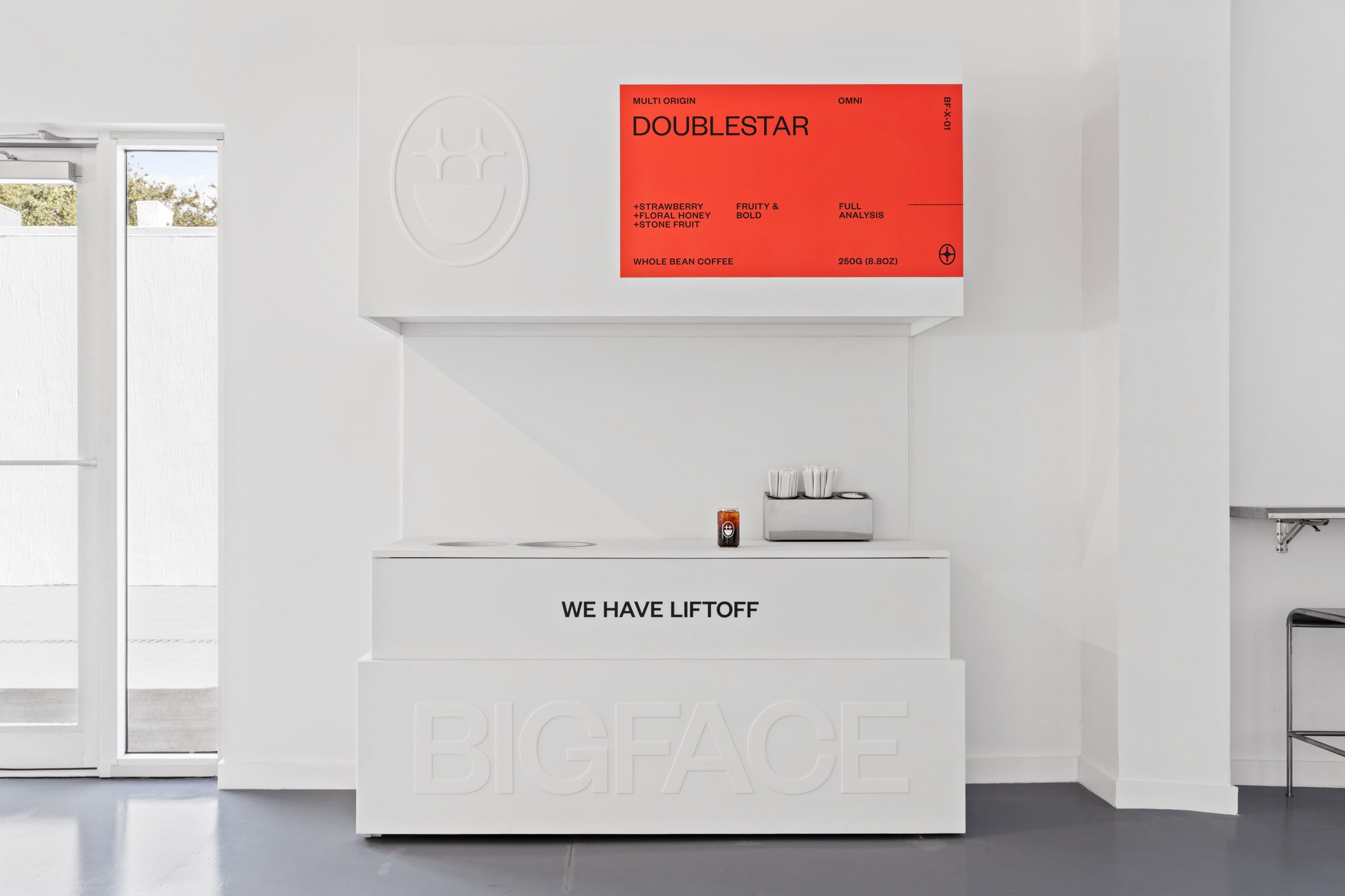 白色牆面搭配立體品牌LOGO雕刻，展現簡約大氣風格。紅色展示牌突顯產品資訊如「Doublestar」咖啡豆的風味特性，強調草莓、花蜜及核果香調。櫃台上的咖啡攪拌棒與蜂蜜瓶設計精巧，注重細節；下方刻有「BIGFACE」字樣的立體字體裝飾，進一步鞏固品牌辨識度
