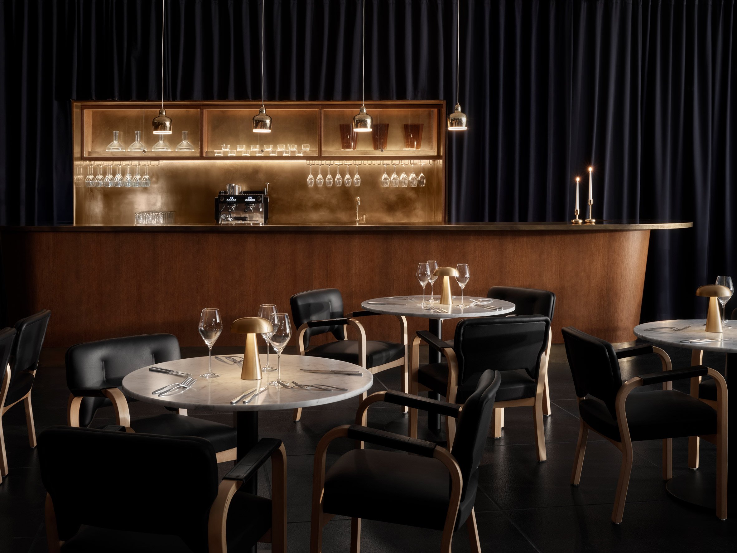 Finlandia Bistro 餐廳:深色布簾與木質吧檯營造沈穩氛圍,金屬背板折射溫暖光澤,圓形石桌與黑色座椅對比鮮明。Artek木扶手椅,突顯用料講究。酒杯與餐具陳列在檯面,讓整個用餐區域更顯迷人