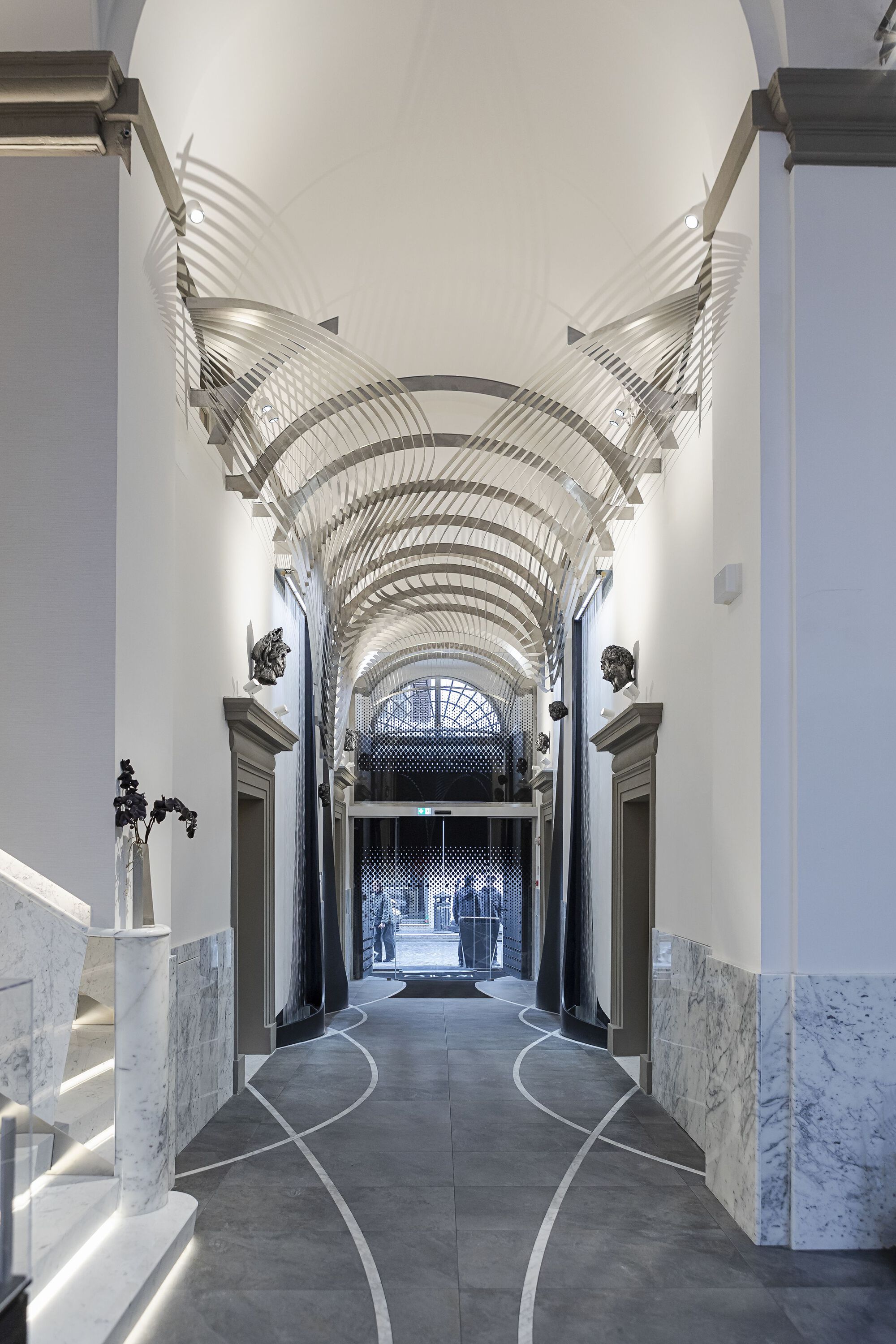 羅密歐酒店(Hotel Romeo Roma)入口通道延續歷史建築的比例,金屬拱形裝置懸浮於天花,光影交錯塑造律動感。大理石階梯與牆面細節呼應文藝復興時期語彙,地坪則以深色石材搭配流線白色鑲嵌線條,引導視覺向外延伸。牆面雕塑與門框細節增添古典氛圍,遠處的金屬格柵入口透出羅馬街景,使室內外形成層次過渡