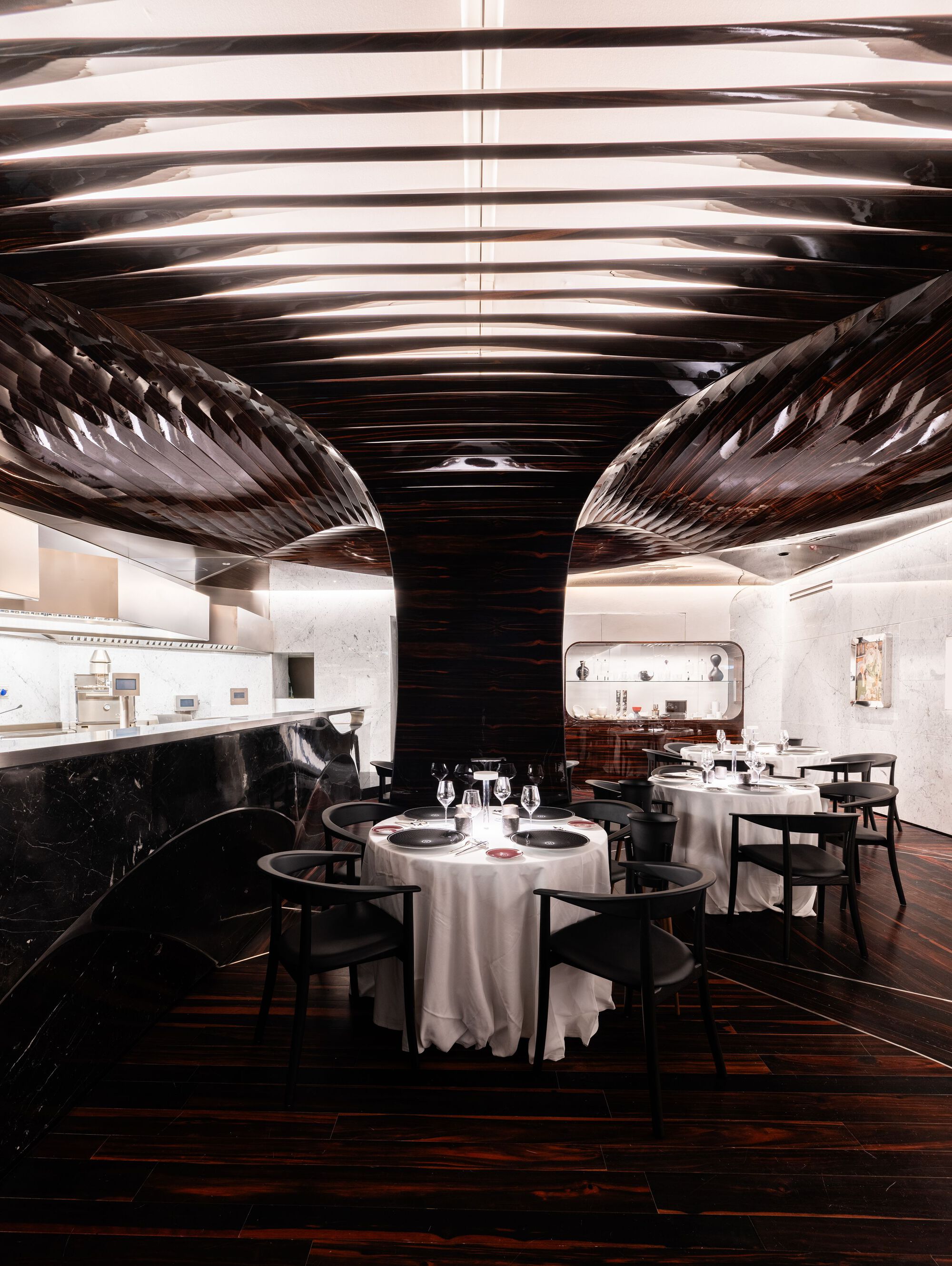 羅密歐酒店（Hotel Romeo Roma）餐廳以拋光黑檀木打造雕塑感天花，流動的線條塑造沉穩氛圍。開放式廚房的黑色大理石吧台延續建築語彙，對比白色桌布與精緻餐具。黑色木椅選用 Carl Hansen & Søn CH88，展現北歐簡約美學。間接照明嵌入天花結構，使光線柔和漫射，強調材質層次。背景展示區採白色大理石襯底，點綴藝術品，使餐飲空間兼具視覺與感官的豐富體驗