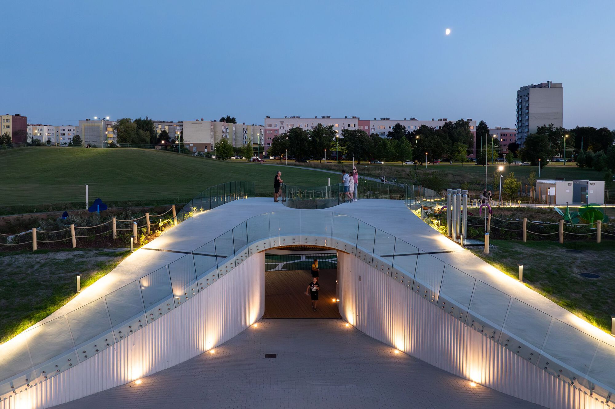 夜晚的Jaworek公園展現溫暖的燈光氛圍,步道以玻璃護欄與曲線設計塑造通透感,下方通道的燈光強調立面的金屬波紋材質,增添現代感