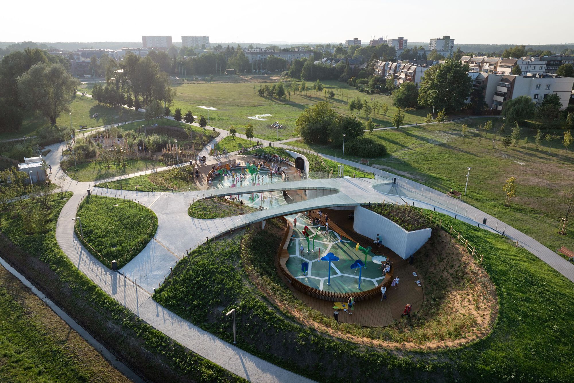 鳥瞰 Jaworek 公園:展示了水樂園區與自然遊戲區的巧妙規劃,蜿蜒步道將空間連結,綠地與人造土丘形成自然屏障。高架通道設有觀景平台,提供遊覽者俯瞰全景的視角。水樂園內的木製座椅與色彩鮮豔的設施相映成趣,打造充滿活力的遊憩空間,自然遊戲區則以天然材料建造遊具,提升孩童的探索與互動體驗