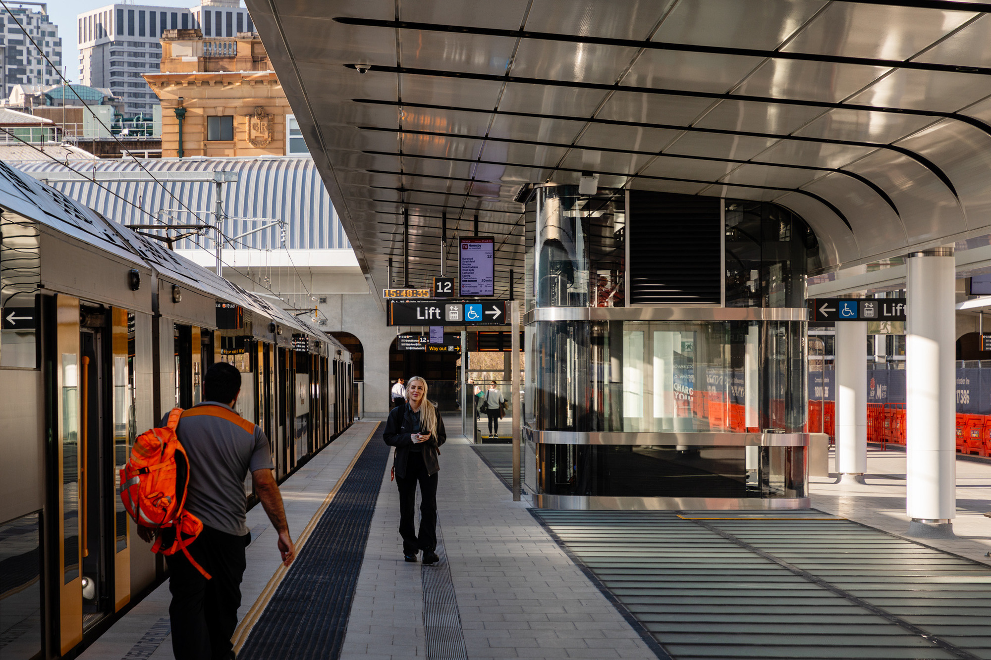 雪梨中央車站 Sydney Central Station Renovation Woods Bagot  改造後的雪梨中央車站月台區展現出現代化設計，採用不鏽鋼與玻璃材質，打造出明亮與清晰的空間。屋頂流線型結構提供遮蔽，並讓自然光透過，提升空間舒適感。新設的電梯與資訊顯示屏優化旅客導引與無障礙設施，展現人性化規劃。背景中的歷史建築與現代結構並存，呈現多元層次的空間語彙