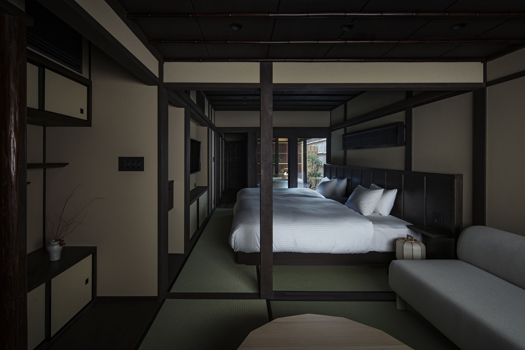 Nazuna Kyoto Higashihonganji 京都 旅館 東本願寺 STUDIO ALUC 深色木構與綠色榻榻米構築條狀動線，床頭延展至壁面，白色圓角沙發與邊几低調安置，開窗望向庭院，室內留白予光影細緻鋪展