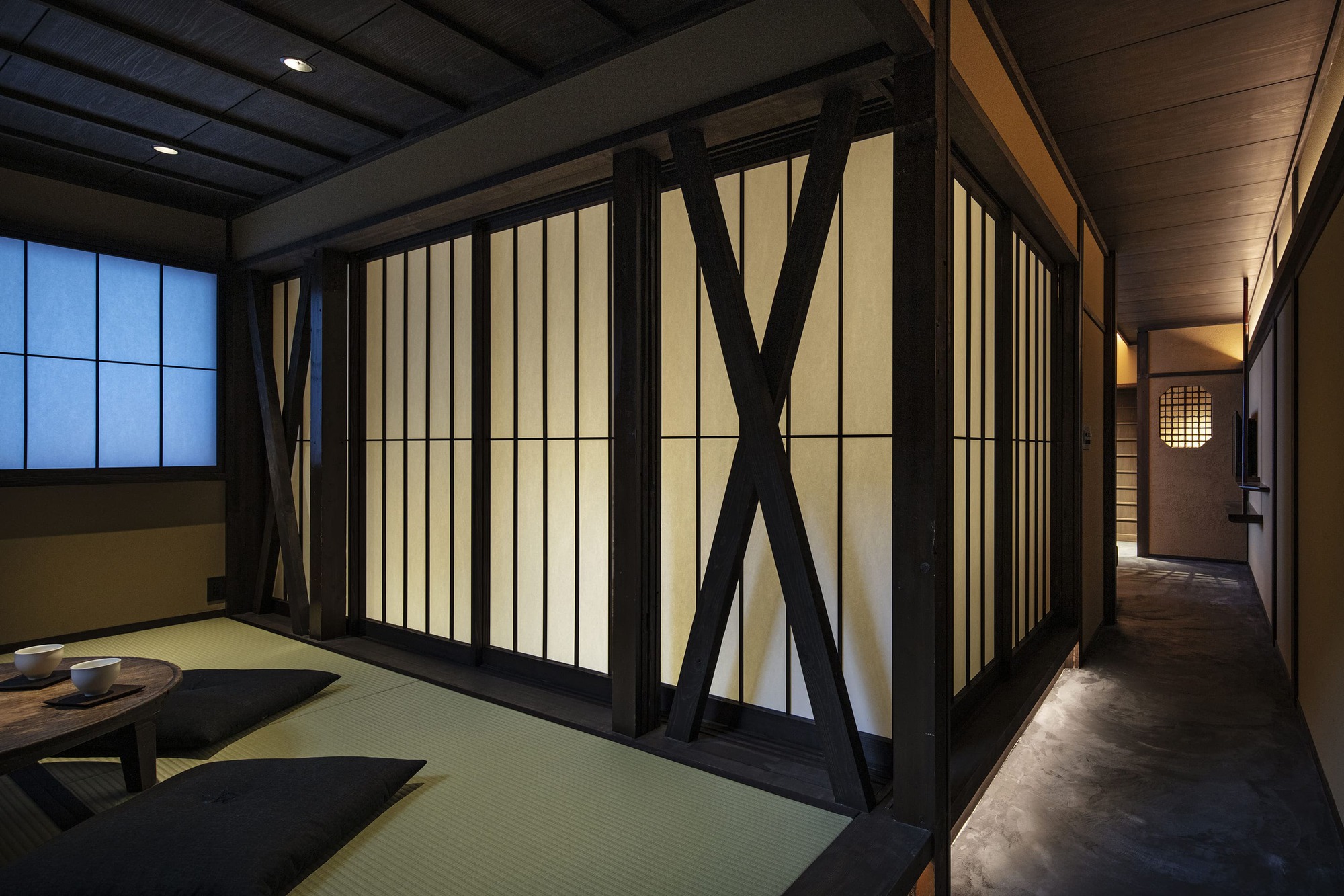 Nazuna Kyoto Higashihonganji 京都 旅館 東本願寺 STUDIO ALUC 榻榻米鋪陳於室內，格子拉門與斜撐構架交錯成景，光線由走廊灑入，圓窗投影牆面，低矮木几與坐墊圍塑親密尺度