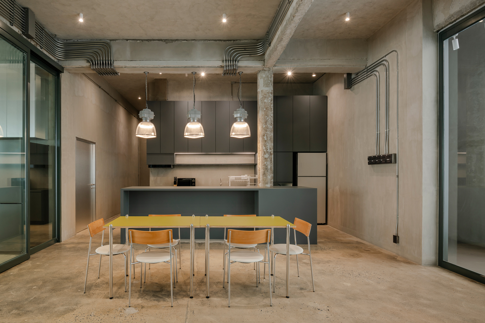 TasteSpace 室內設計 泰國曼谷冠軍咖啡店「Factory Coffee」「Factory Coffee」總部二樓被規劃為工作坊和辦公室，工業吊燈與裸露管線強化原始氛圍，中央吧台結合灰色收納櫃與微波爐，簡潔長桌及木椅則提供輕鬆用餐與討論區域