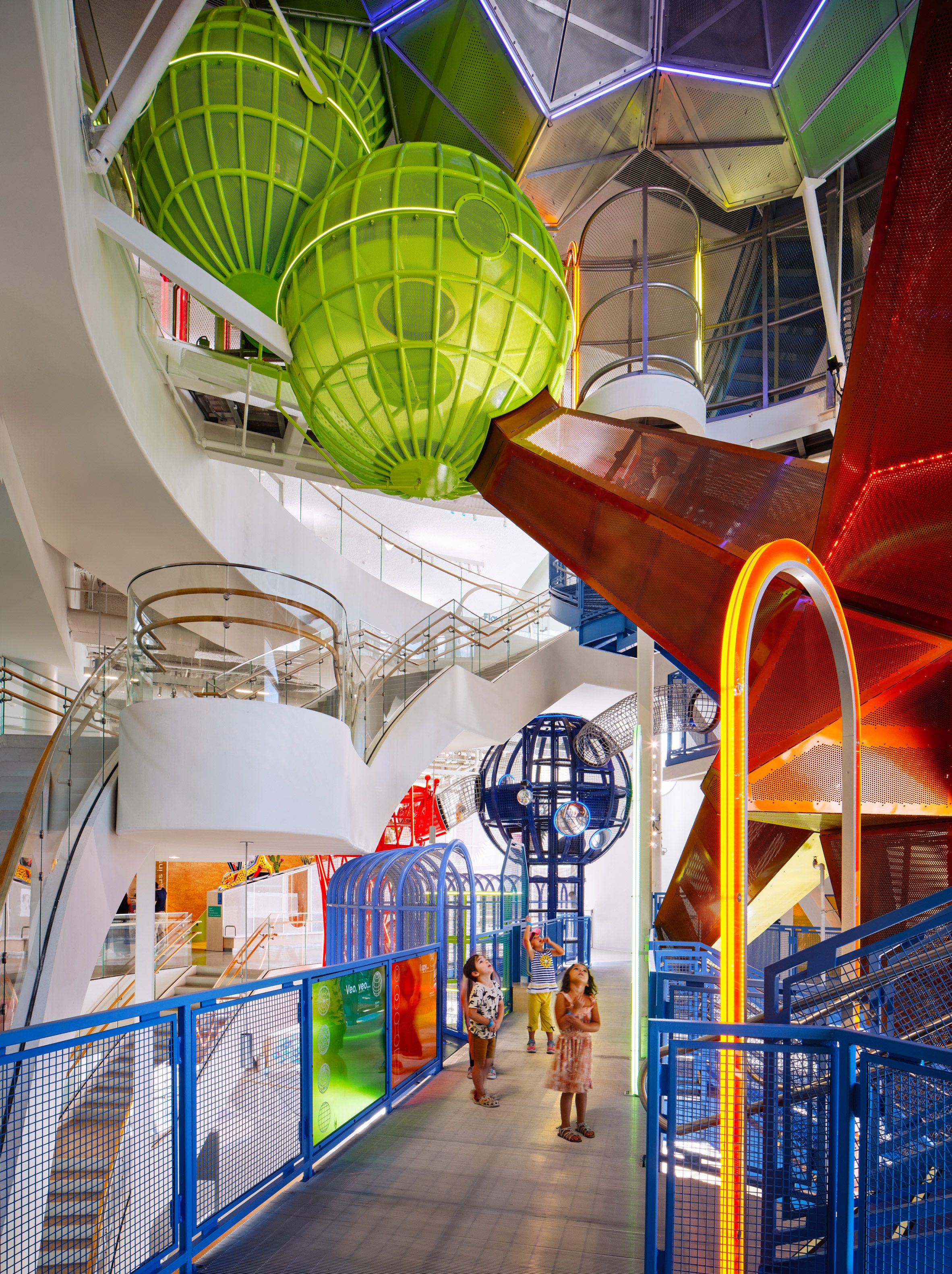 La Nube 兒童中心內部，懸掛的綠色與藍色球體裝置錯落於多層空間，提供攀爬與探索的遊樂體驗。蜿蜒的白色樓梯與玻璃欄杆勾勒出流動動線，金屬網格橋樑與沖孔鋼板滑梯串聯不同樓層，形成立體遊戲路徑