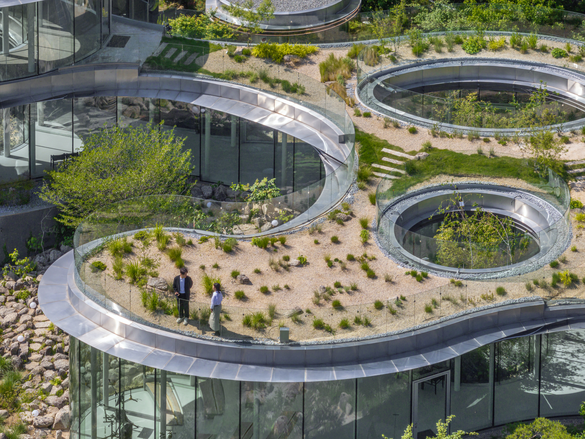 韓國複合文化空間蔚山市蔚州郡「MIGIUI」Architect-K 起伏屋頂覆以砂礫與耐旱植栽,曲線邊界對應地形弧度,形塑高低錯落的漫遊平台。圓形天井開口留住樹冠與天光,為下方空間引入垂直景深,整體如同地景再造的另一層呼吸