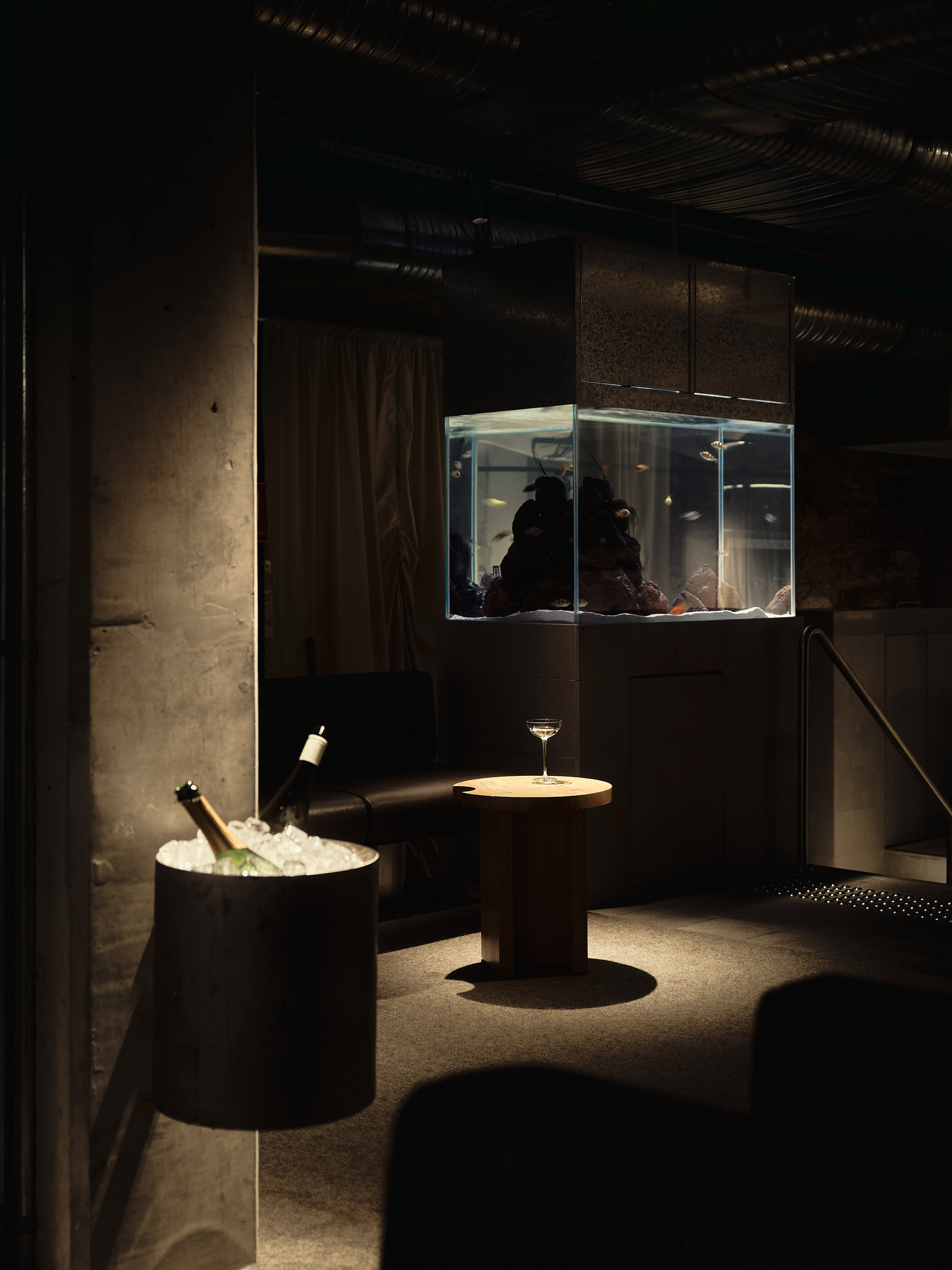 低矮天花佈滿管線，水族箱懸掛空中成為微型舞台，原木小桌與嵌牆冰桶構築出暫停節奏的深夜社交節點
