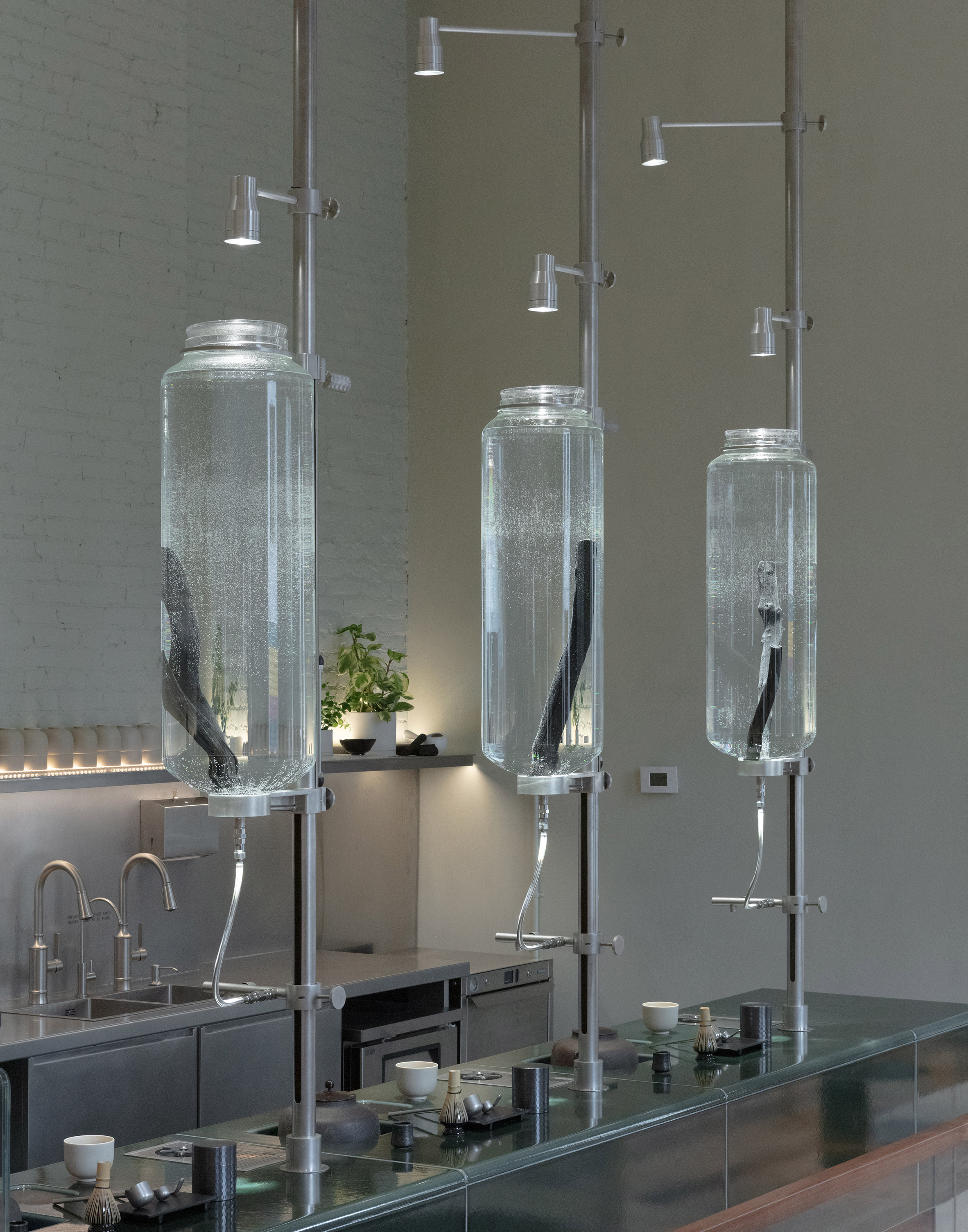 吧台後方三座大型玻璃容器昂然佇立，浸泡著數根碩大的日本備長炭（Binchotan）。這套由 Ciguë 為永續辦公環境開發的過濾系統，在此被重新設計，以滿足12 Matcha的商業營運需求