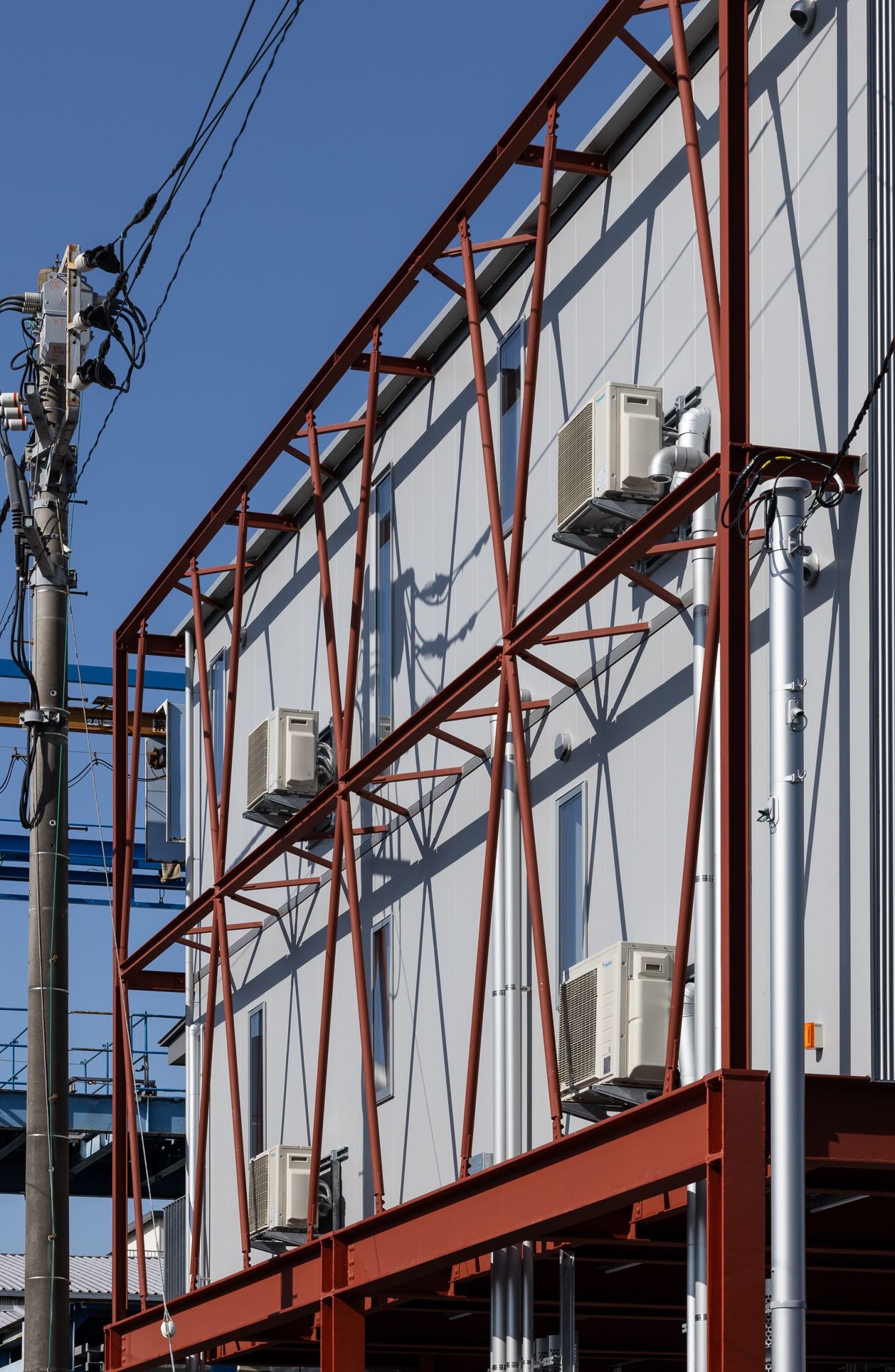 野口修 Sanmei 辦公室 建築東側立面展現獨特構造工法。鮮紅色的鋼骨與斜撐，刻意從主牆面脫開70公分，形成一道垂直的維護通道。所有的空調室外機與管線均被整合於此，便於日後檢修，也維持了主要牆面的完整性