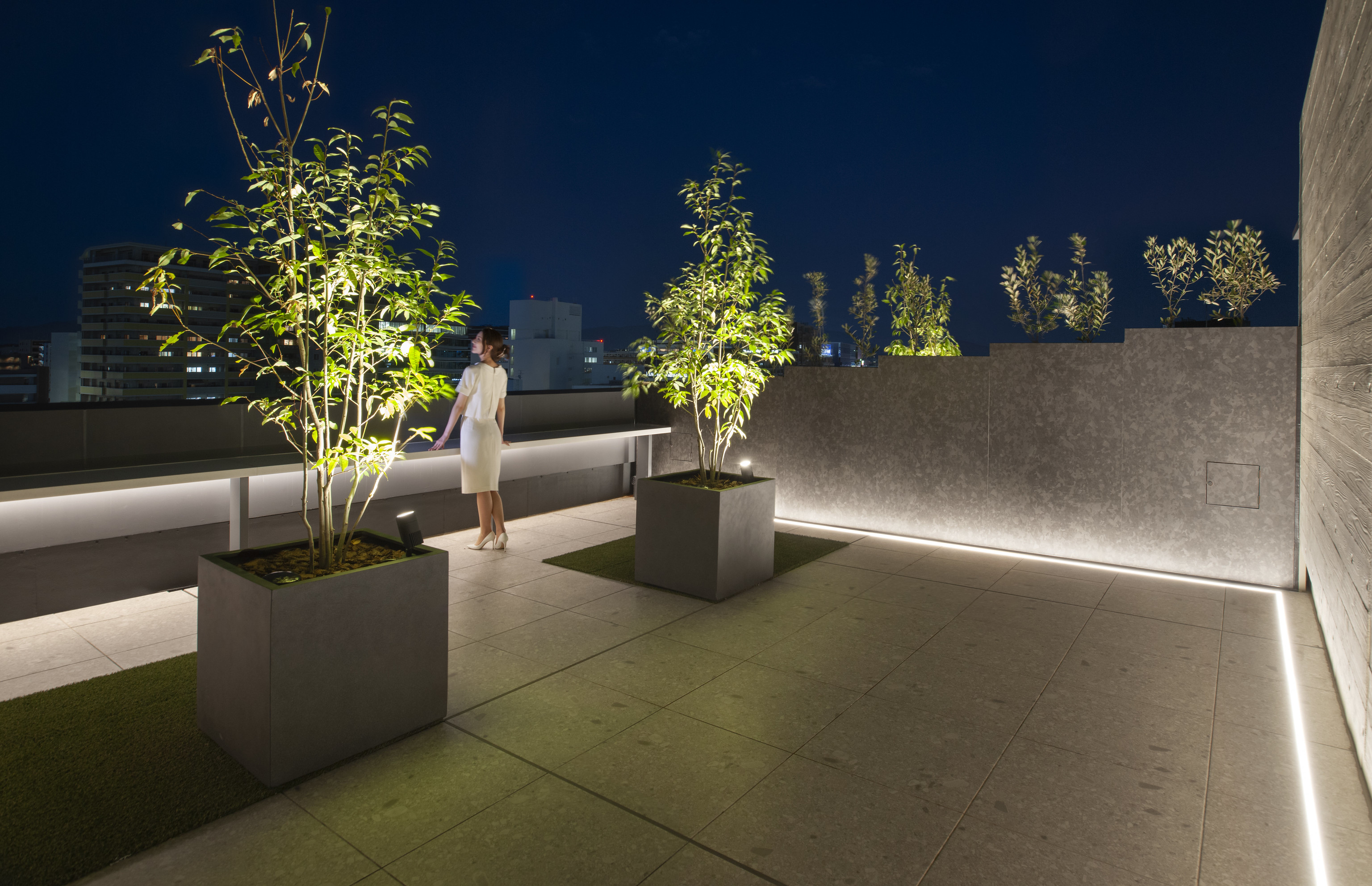 迫慶一郎 Keiichiro Sako SAKO建築設計工場 SAKO Architects 福岡 VECTORS in Fukuoka 辦公大樓 屋頂露臺以低照度燈光與植栽為界，鋪面選用石材模矩拼貼，呈現沈穩質地。俯瞰博多灣天際，都市光點與綠意交錯，成為商辦環境中一處夜間喘息的開放節點