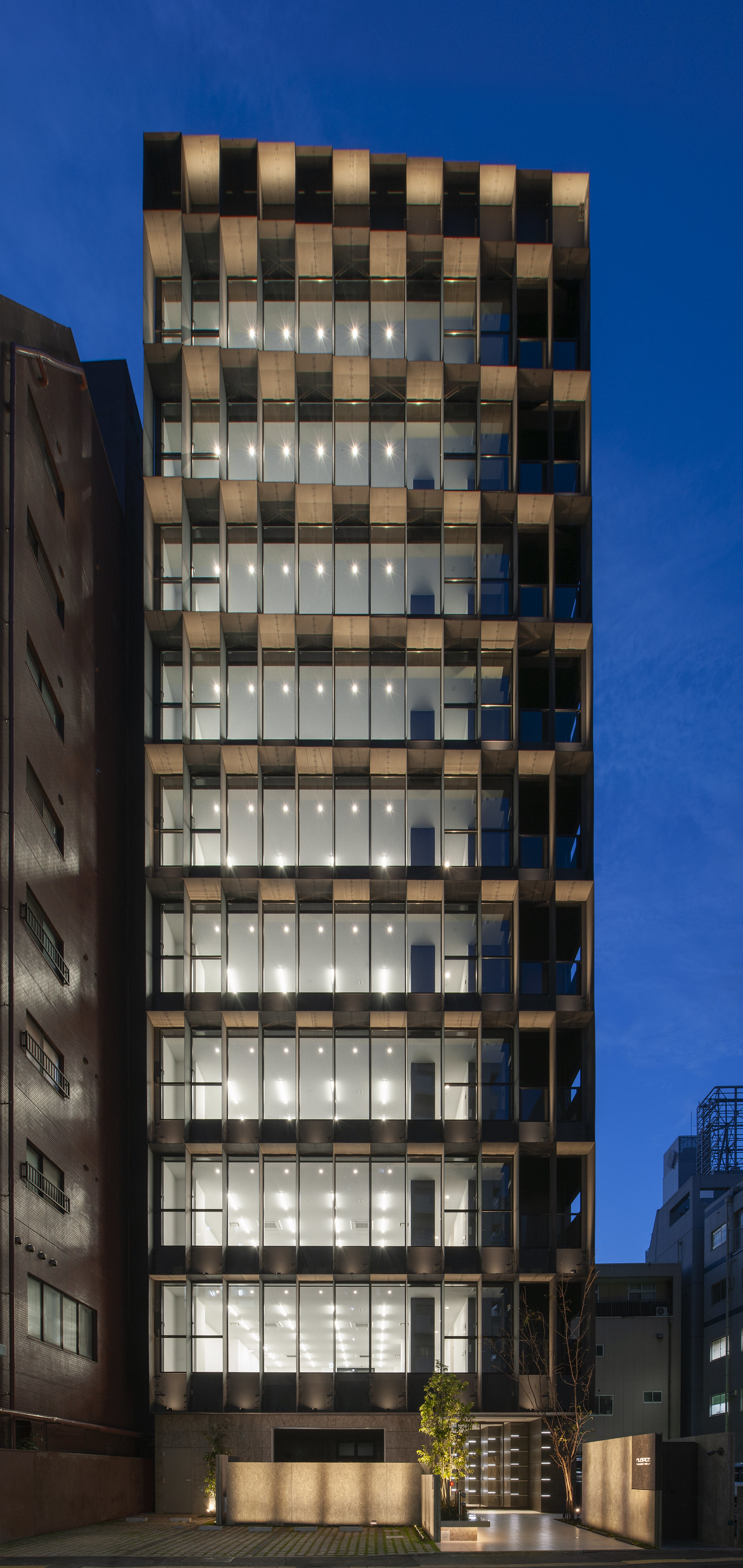 迫慶一郎 Keiichiro Sako SAKO建築設計工場 SAKO Architects 福岡 VECTORS in Fukuoka 辦公大樓 立面格柵以鋼構縱橫編織，序列與光源在夜色中同步演出。正立面呈現高度秩序的幾何節奏，在兩側高樓夾擊間自成景框