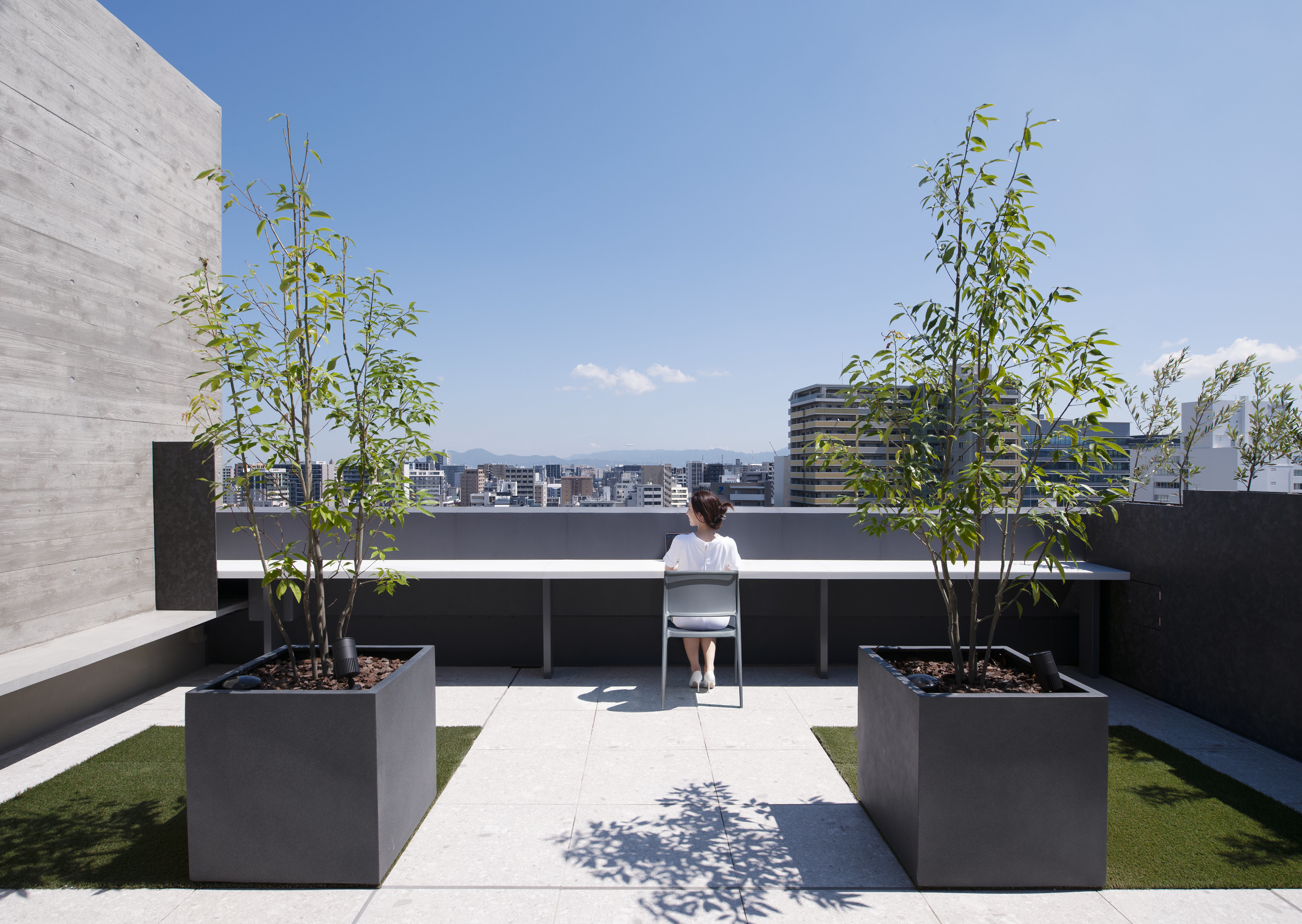 迫慶一郎 Keiichiro Sako SAKO建築設計工場 SAKO Architects 福岡 VECTORS in Fukuoka 辦公大樓 屋頂露臺設置長桌與簡潔座椅，提供俯瞰福岡市景的靜謐端點。石材鋪面與清水混凝土牆面對比柔軟綠意，在高密度都市紋理中開闢一處可停留、可觀景的輕盈轉場