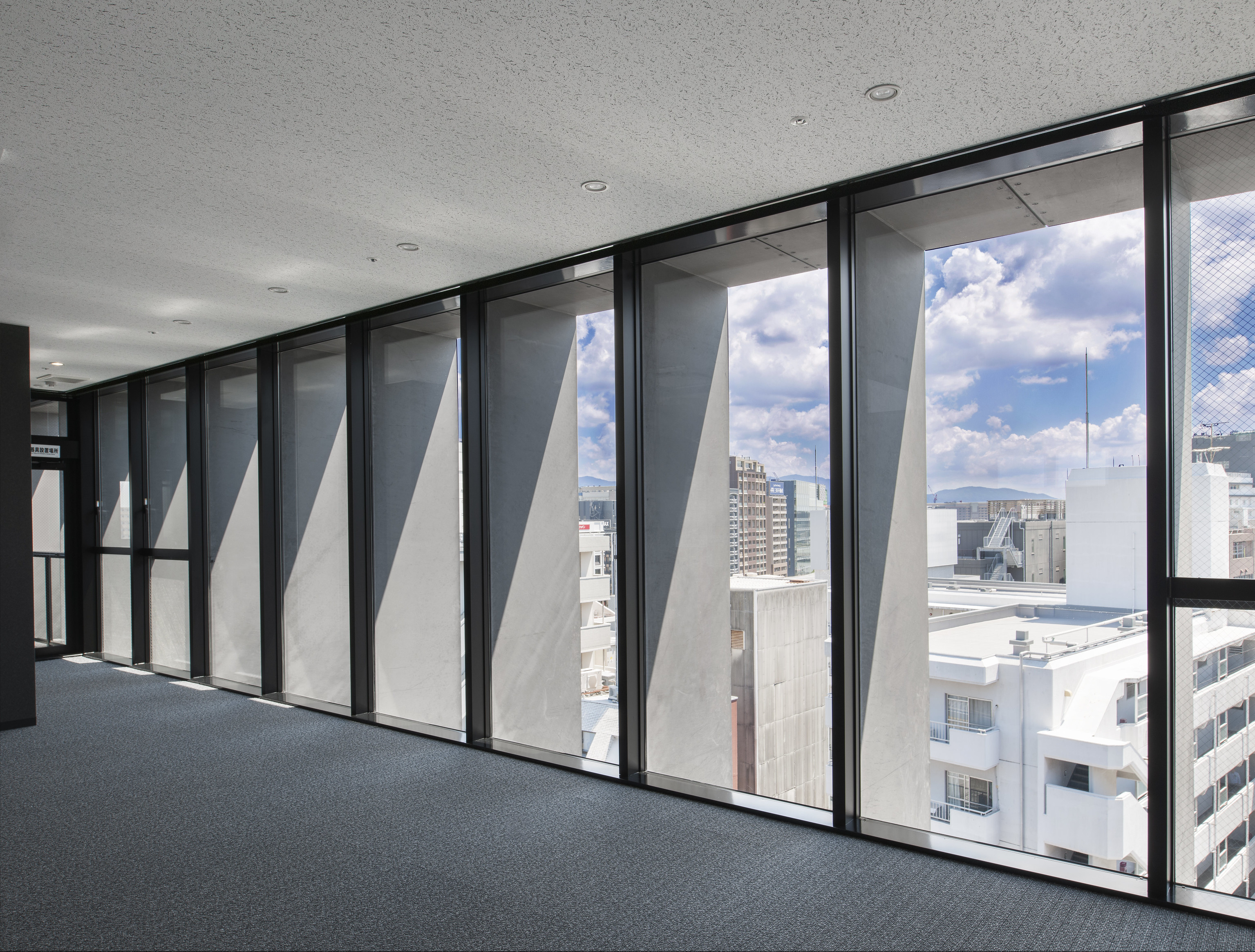 迫慶一郎 Keiichiro Sako SAKO建築設計工場 SAKO Architects 福岡 VECTORS in Fukuoka 辦公大樓 光沿鋼板格柵斜面切入室內，投下如刻度般節奏清晰的陰影，界定出一片無柱的開放工作場域。外部城市輪廓隱約滲入，讓建築與都市景觀維持靜默而連續的對望關係