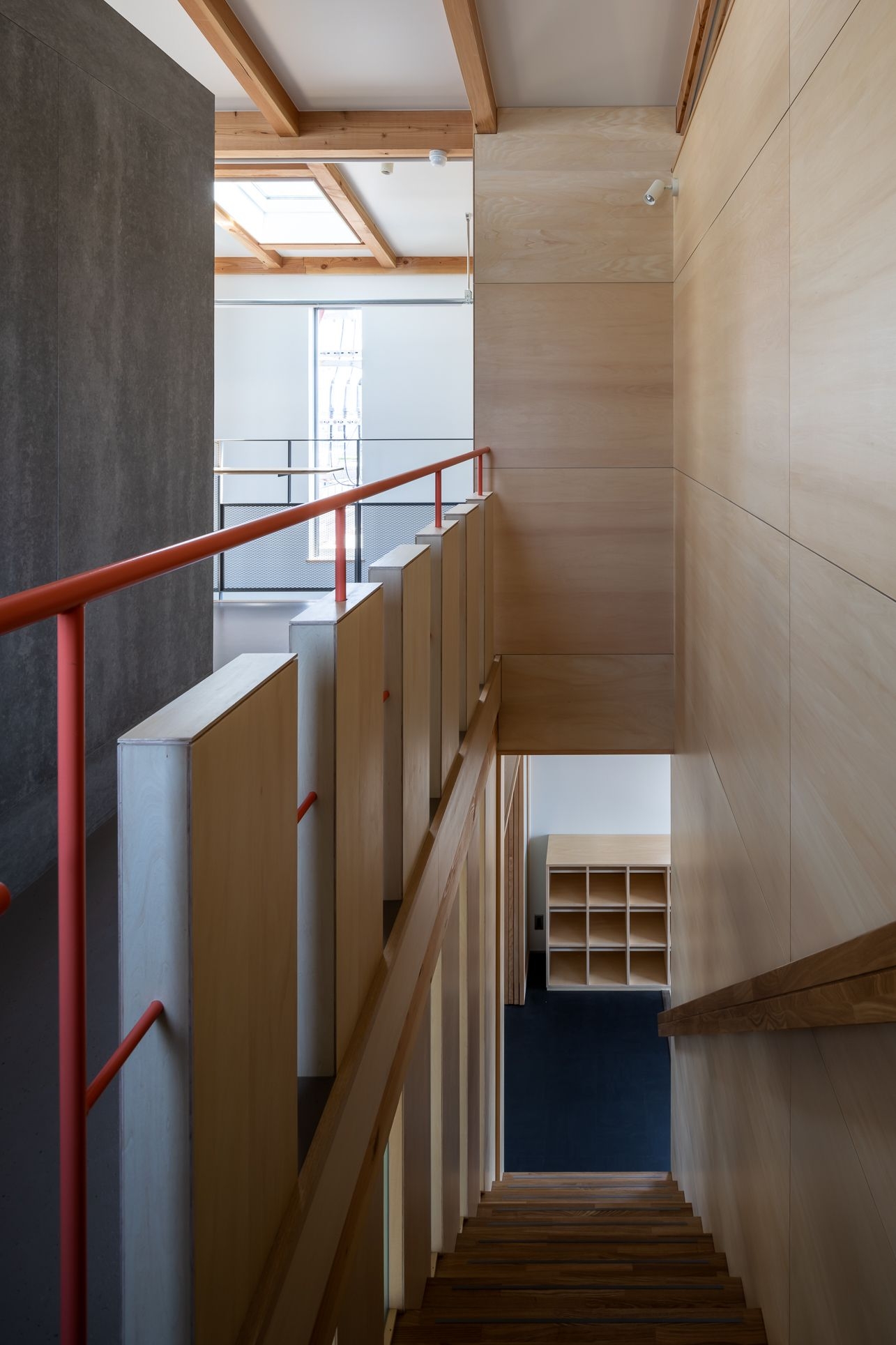 野口修 Sanmei 辦公室 樓梯間是整棟建築的垂直樞紐，集結了此案的關鍵設計語彙。溫潤的木質階梯與夾板牆面，對比著深灰色的壁板；鮮紅色的扶手則化為一道引導動線的視覺彩帶，串起不同樓層
