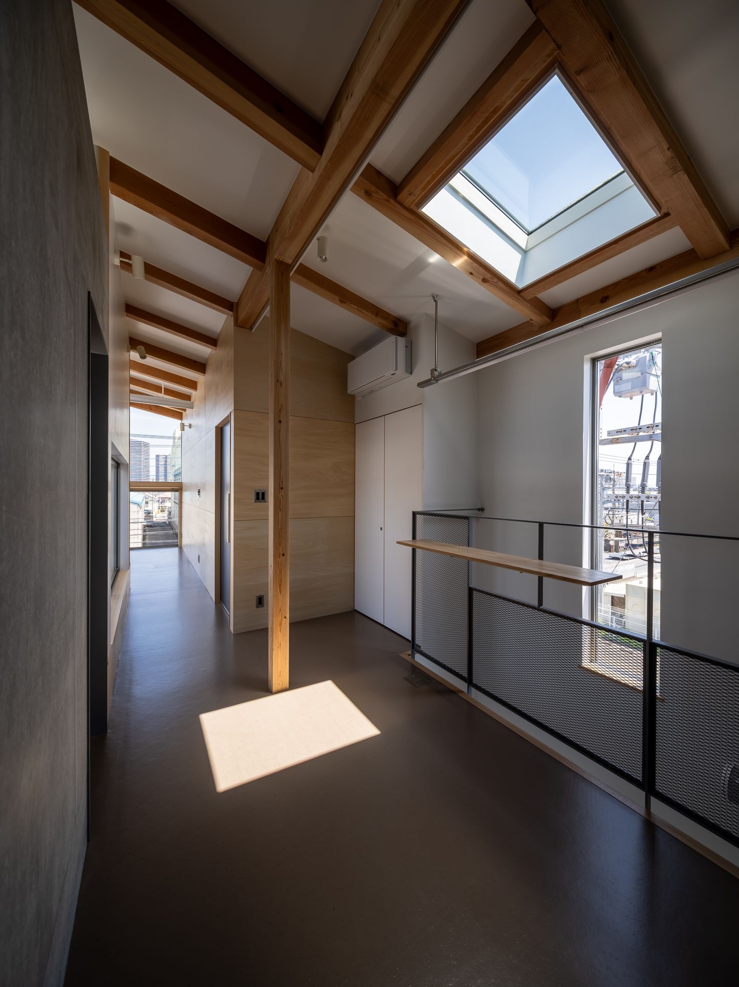 野口修 Sanmei 辦公室 三樓廊道空間，將木構造的魅力展露無遺。厚實的木樑、木柱與桁架，在傾斜的屋頂下構成空間的主要韻律。陽光自天窗灑落，在深色地板上形成溫暖光斑。欄杆採用黑色金屬網搭配木質層板，通透的工業質感，與樸實的木構造、夾板牆面共同圍塑出長屋巷弄般的靜謐氛圍