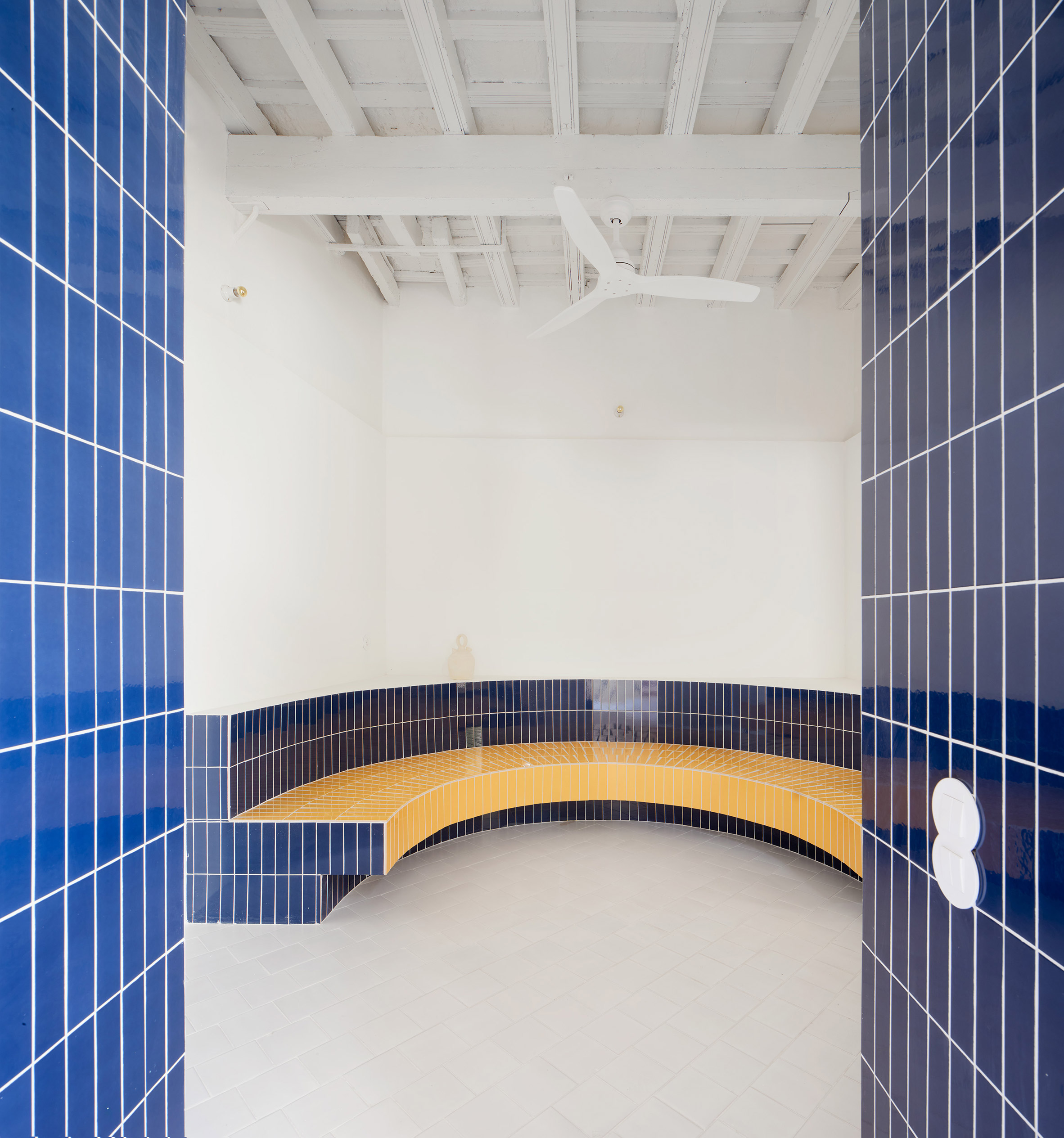 El Puerto de Santa María 住宅室內設計 Lucas y Hernández-Gil 西班牙 此處是對古羅馬浴場更衣室（apodyterium）的當代回望。搶眼的寶藍與檸檬黃磁磚，構成一道大型弧形長凳，成為純白空間中的絕對主角。上方保留了粉刷過的歷史木樑天花板，新舊元素的強烈對比，鋪陳出一種既古典又前衛的空間張力，大膽而充滿詩意