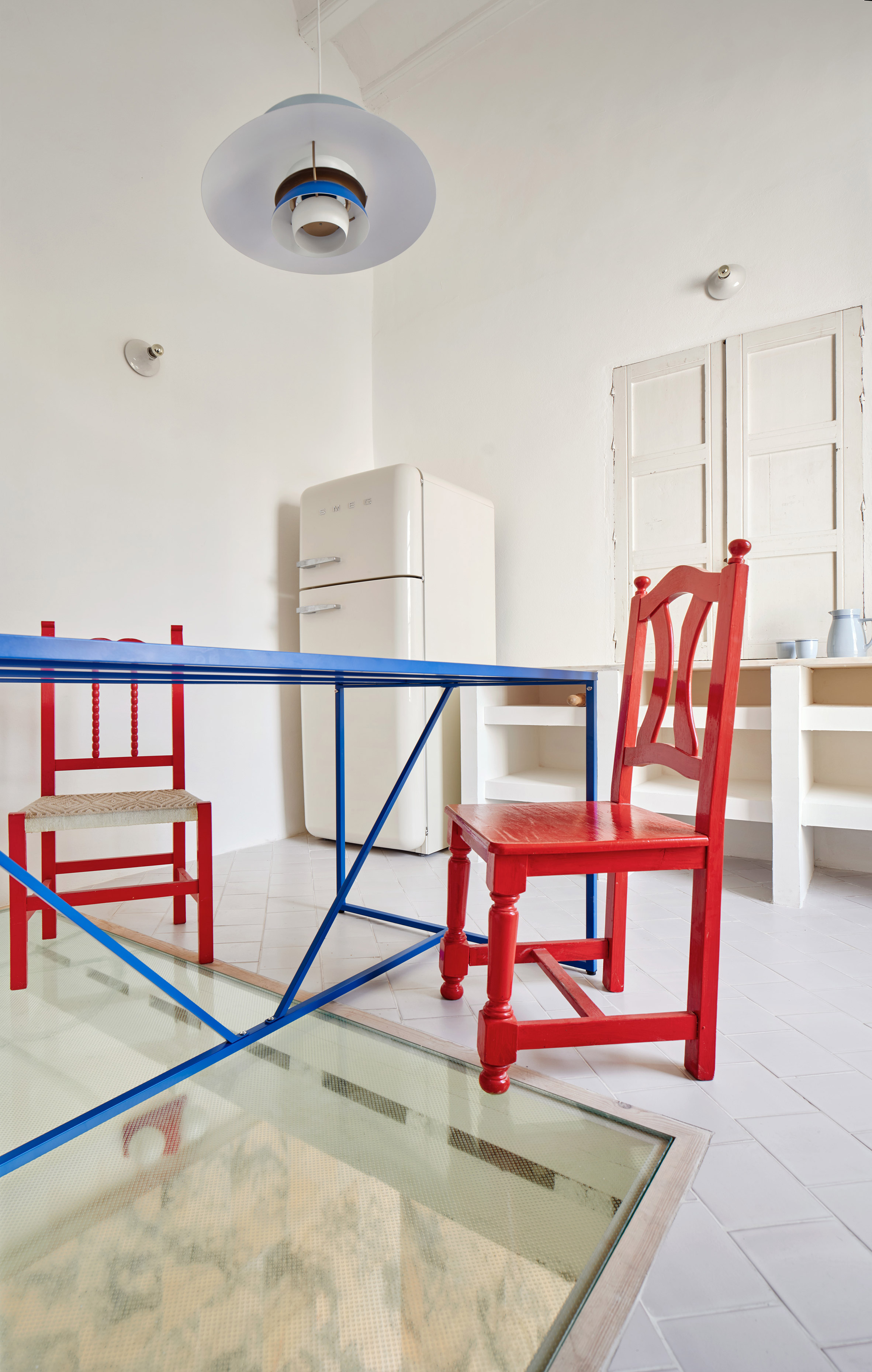 El Puerto de Santa María 住宅室內設計 Lucas y Hernández-Gil 西班牙 二樓廚房展現了遊戲般的精神，大片透明玻璃地板讓樓下泳池水光盡收眼底。傳統木椅被賦予奔放的鮮紅，對比著寶藍色桌腳，與上方 Poul Henningsen 設計的PH 5經典吊燈，共同構成一幅跨越時代的設計拼貼畫