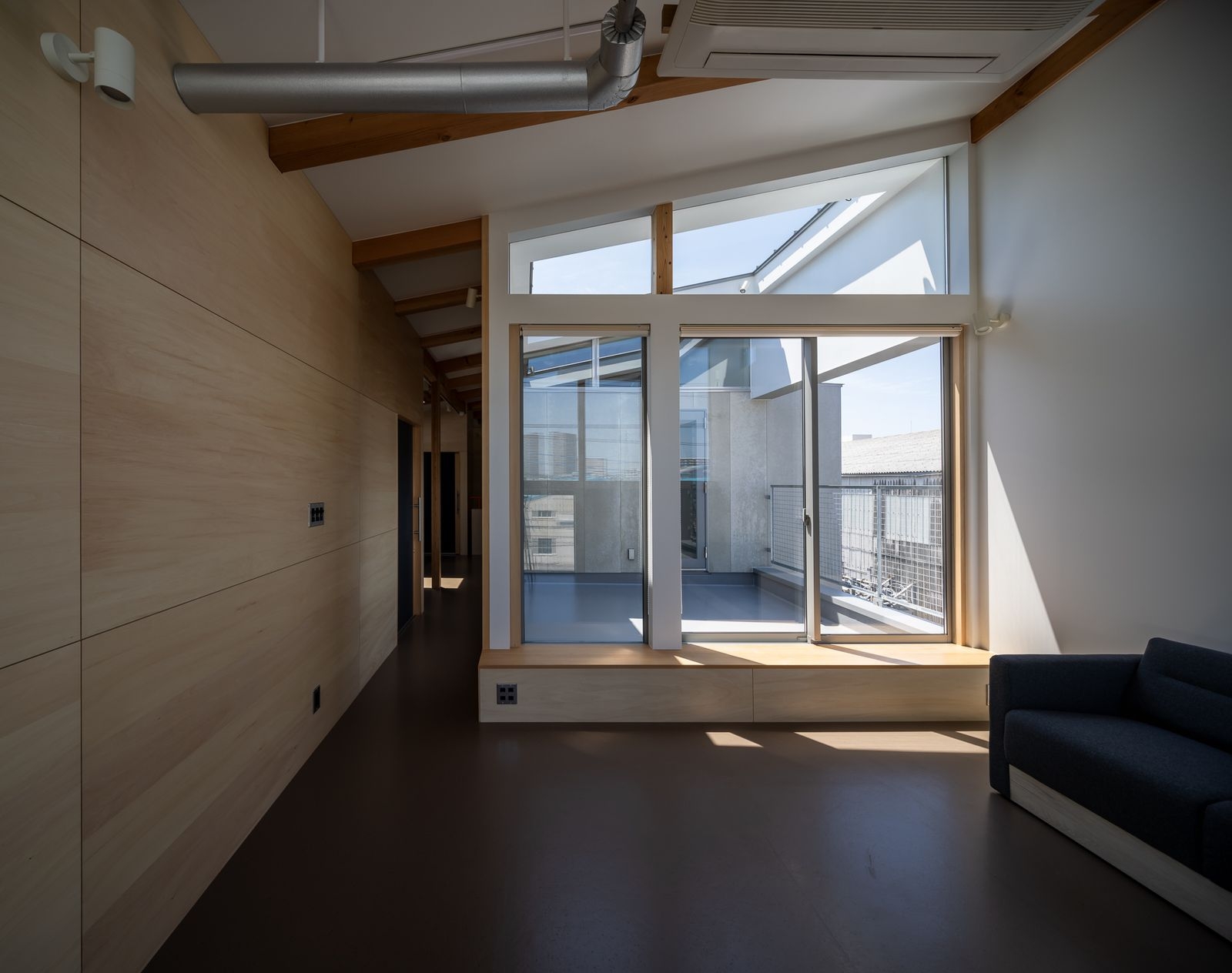 野口修 Sanmei 辦公室 三樓的休憩區，清楚演繹了「長屋」的巷弄（路地）概念。一道長廊串連起各個單元，一側則以大面開窗迎向內縮的私密陽台。傾斜的屋頂下，外露的木樑、管線與溫潤的夾板牆面共同定義空間調性。窗下的臥榻式收納櫃，則為此處增添了濃厚的起居氣息