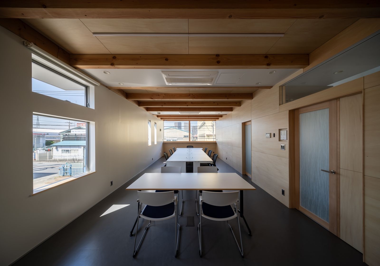 野口修 Sanmei 辦公室 辦公室呈現為一道長廊般的開闊空間，天花板大方地展露木造結構的序列感。溫潤的木質膠合樑、夾板牆面與天花，和深色的地板形成對比。家具選用基礎的會議桌椅，維持空間的純粹。窗戶引入了光線與周遭的工業地景