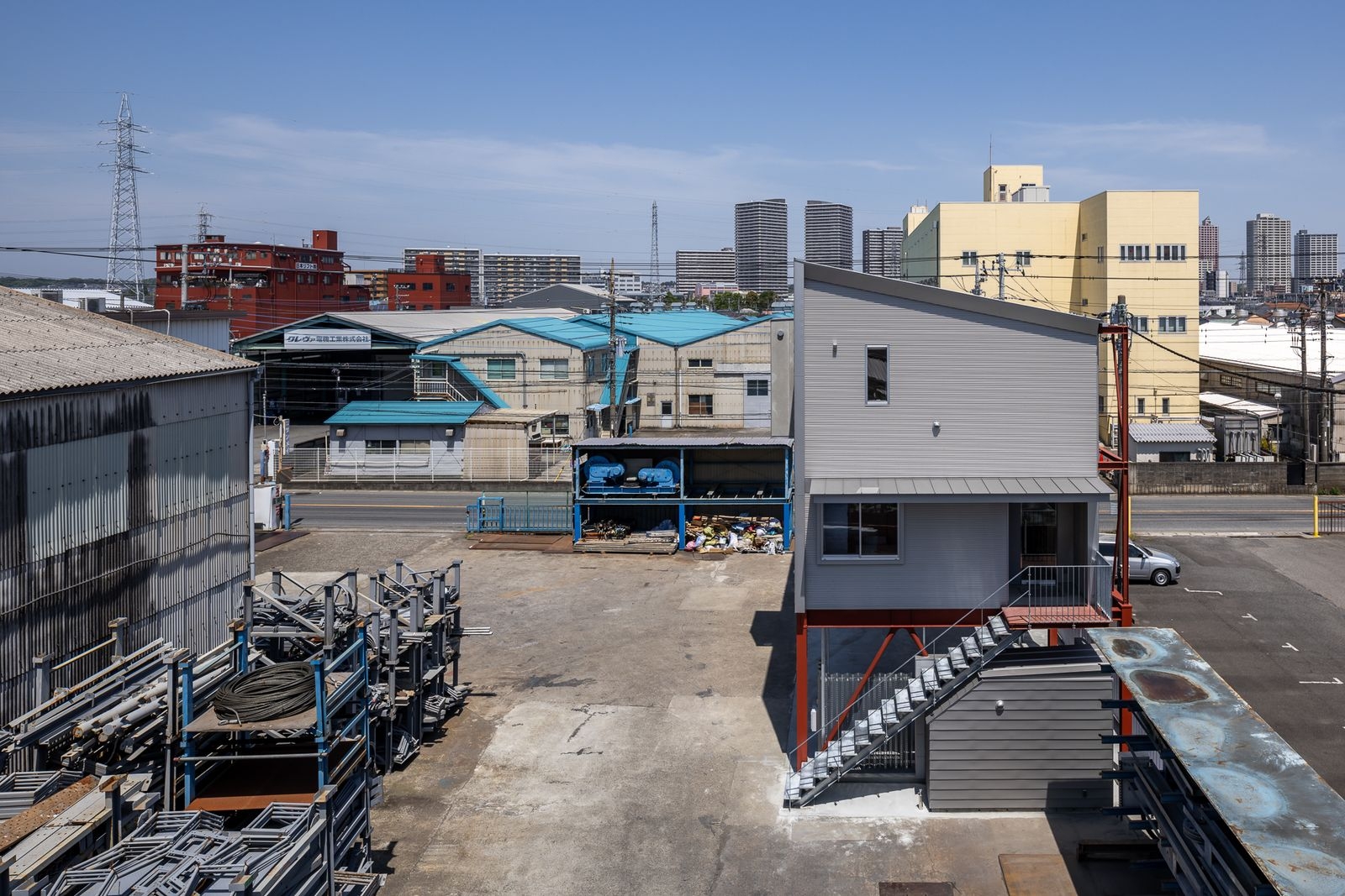 略顯雜亂的機能性場域裡，這座有著單斜屋頂的灰色量體，以其清晰的幾何造型與秩序感，成為一個沉靜而自持的存在。日本建築師野口修並未選用昂貴的材料去對抗環境，而是將鍍鋅鋼梯、金屬壁板等工業語彙重新編排，透過設計賦予它們嶄新的價值