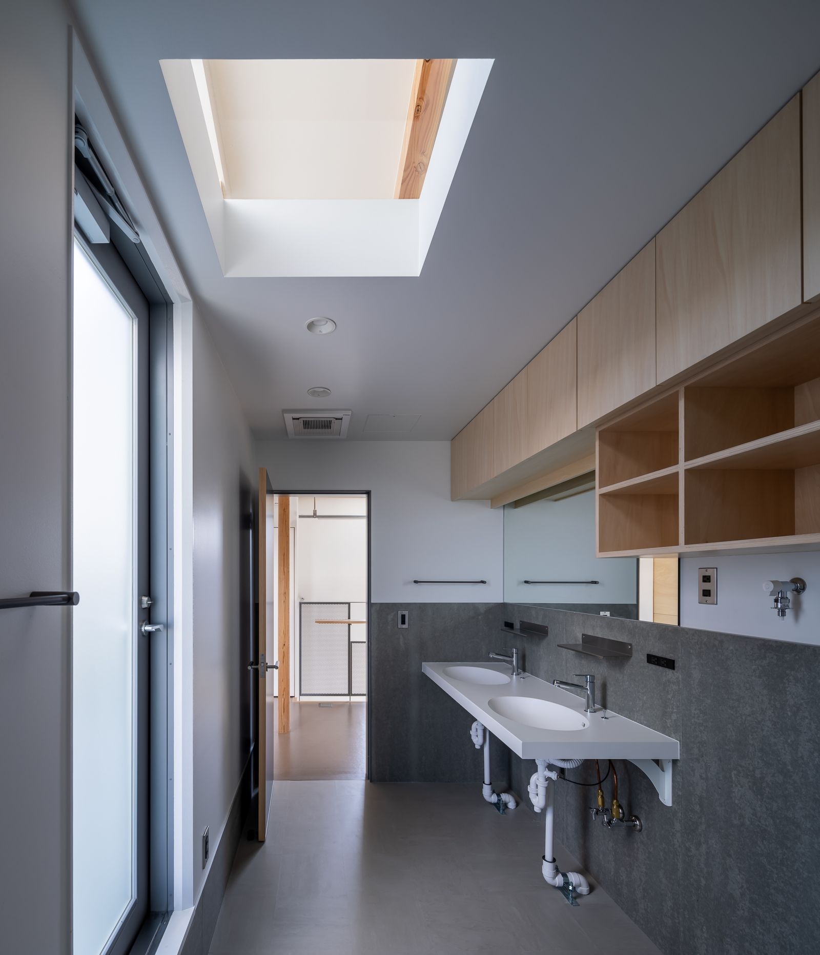野口修 Sanmei 辦公室 洗面室（洗手間）的設計，將「道具立」的務實精神展露無遺。天窗引入自然光，照亮了由夾板、深色壁板構成的線性空間。水槽下方的管線特意不做遮掩，直接裸露，將維修的便利性置於首位。這種直白的設計手法，從公共空間一路延伸至後勤場域，貫徹了整棟建築的設計主張