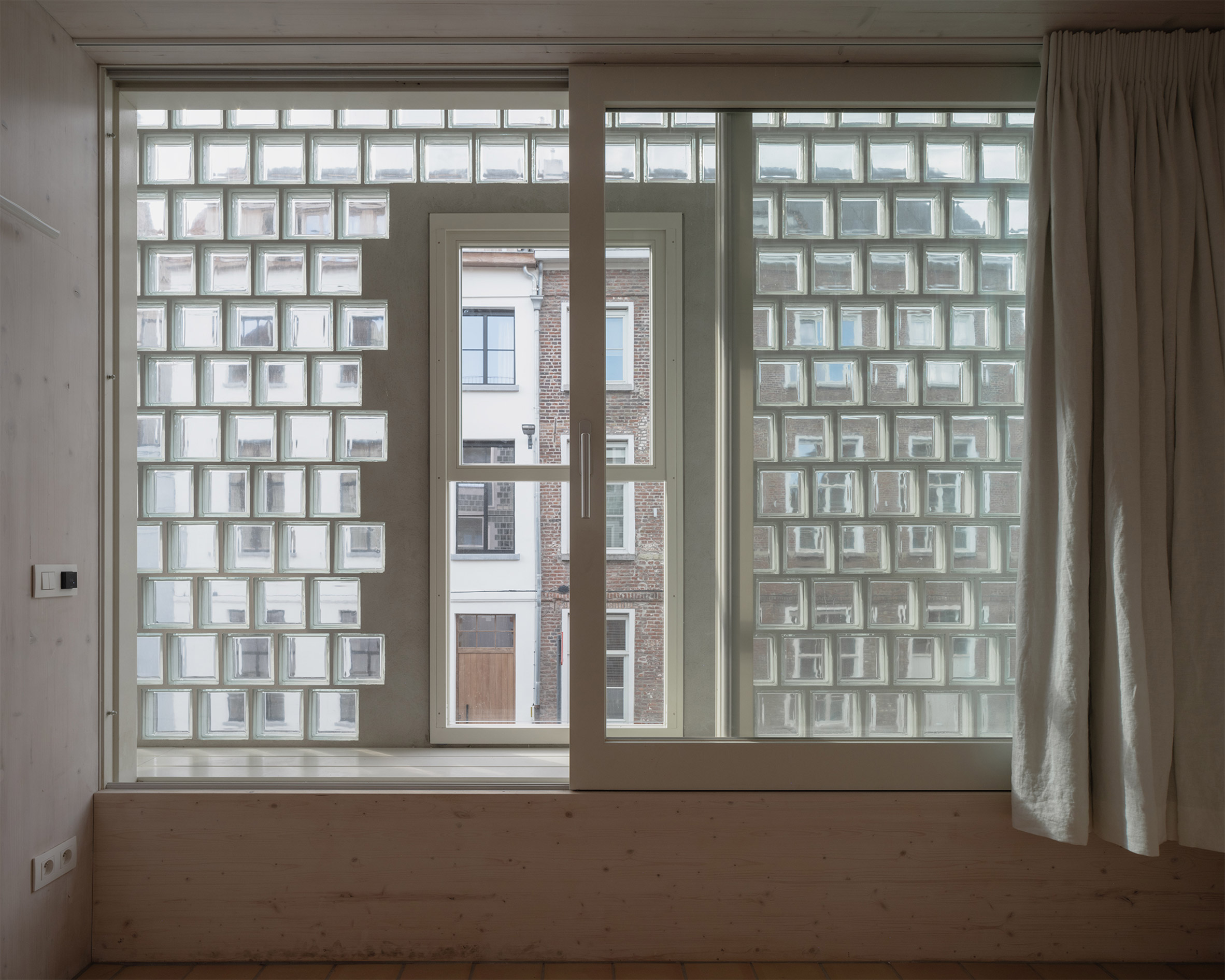 比利時 根特 Ghent 玻璃磚屋 Delmulle Delmulle Architecten Glass Brick House 光線穿透雙層玻璃磚牆灑入室內，模糊街景輪廓，保留隱私同時引入通透感。原木窗框與CLT牆體質樸相應，窗台座位與亞麻窗簾勾勒出一處安靜凝視城市的日常所在