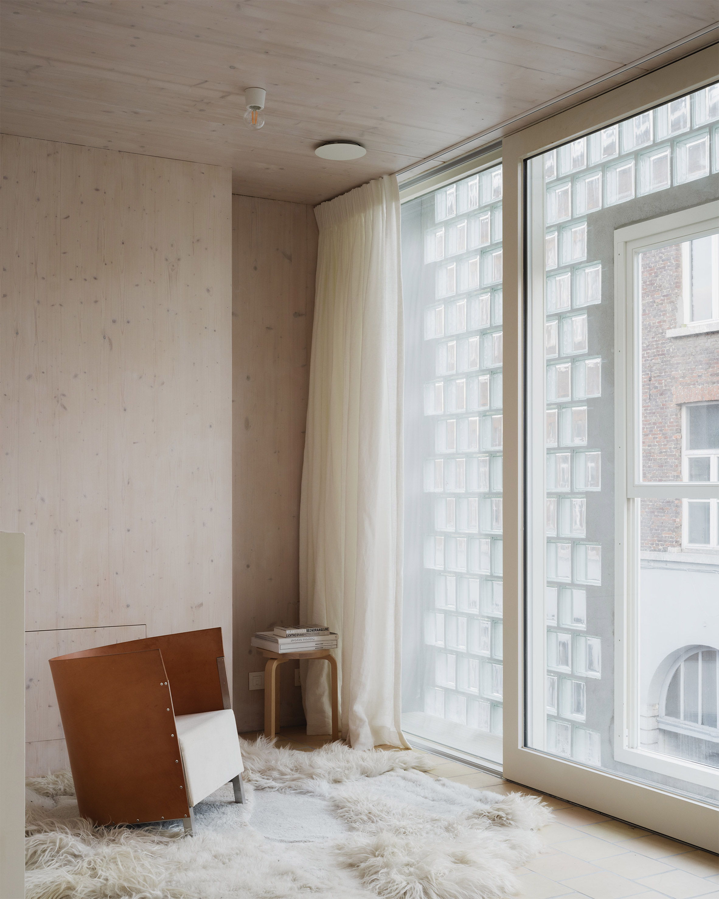 比利時 根特 Ghent 玻璃磚屋 Delmulle Delmulle Architecten Glass Brick House 透過玻璃磚濾入的柔光灑落CLT木牆，形成寧靜角落。Alvar Aalto 經典茶几搭配皮革單椅，置於羊毛地毯之上，簡約中流露溫度，成為高密度城市中稀有的靜謐居所