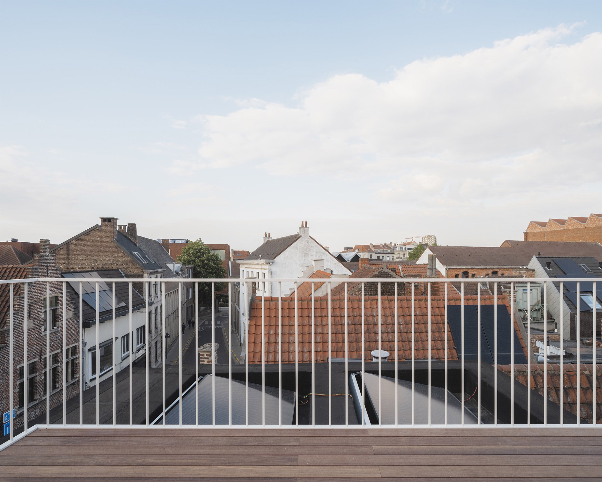 比利時 根特 Ghent 玻璃磚屋 Delmulle Delmulle Architecten Glass Brick House 屋頂露臺鋪設溫潤木地板，纖細白色欄杆輕輕框住根特老城街景。此處遠離街面擁擠，俯瞰磚瓦屋脊與天光雲影，是在密集城市中保留的一段私密呼吸縫隙