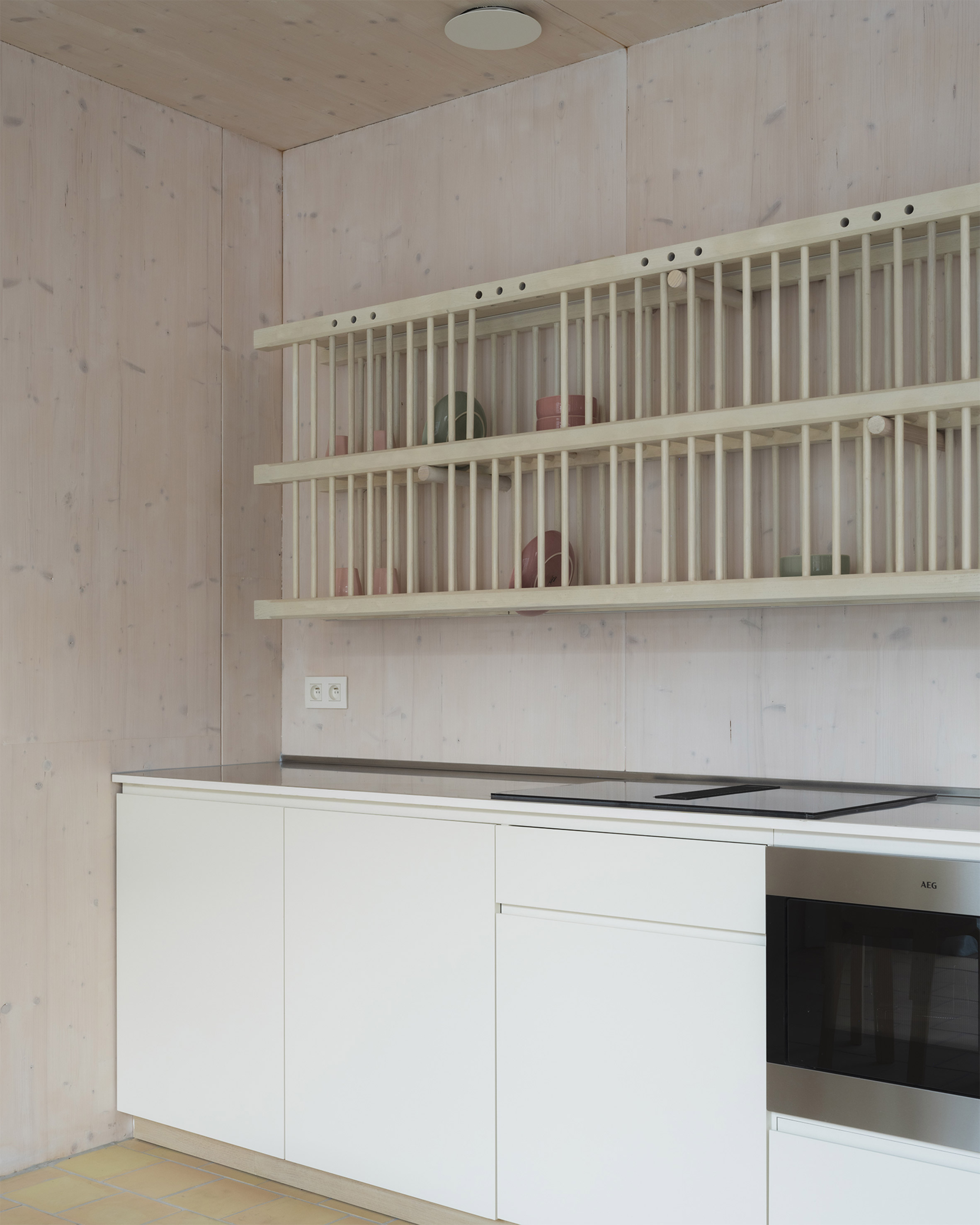 比利時 根特 Ghent 玻璃磚屋 Delmulle Delmulle Architecten Glass Brick House 簡潔白色廚櫃搭配AEG嵌入式爐具，烘托CLT木牆的溫潤質地。開放式木製碗架以細桿構成節奏感，陶器陳列其間，構築出在極簡與手感之間輕聲低語的日常廚房風景