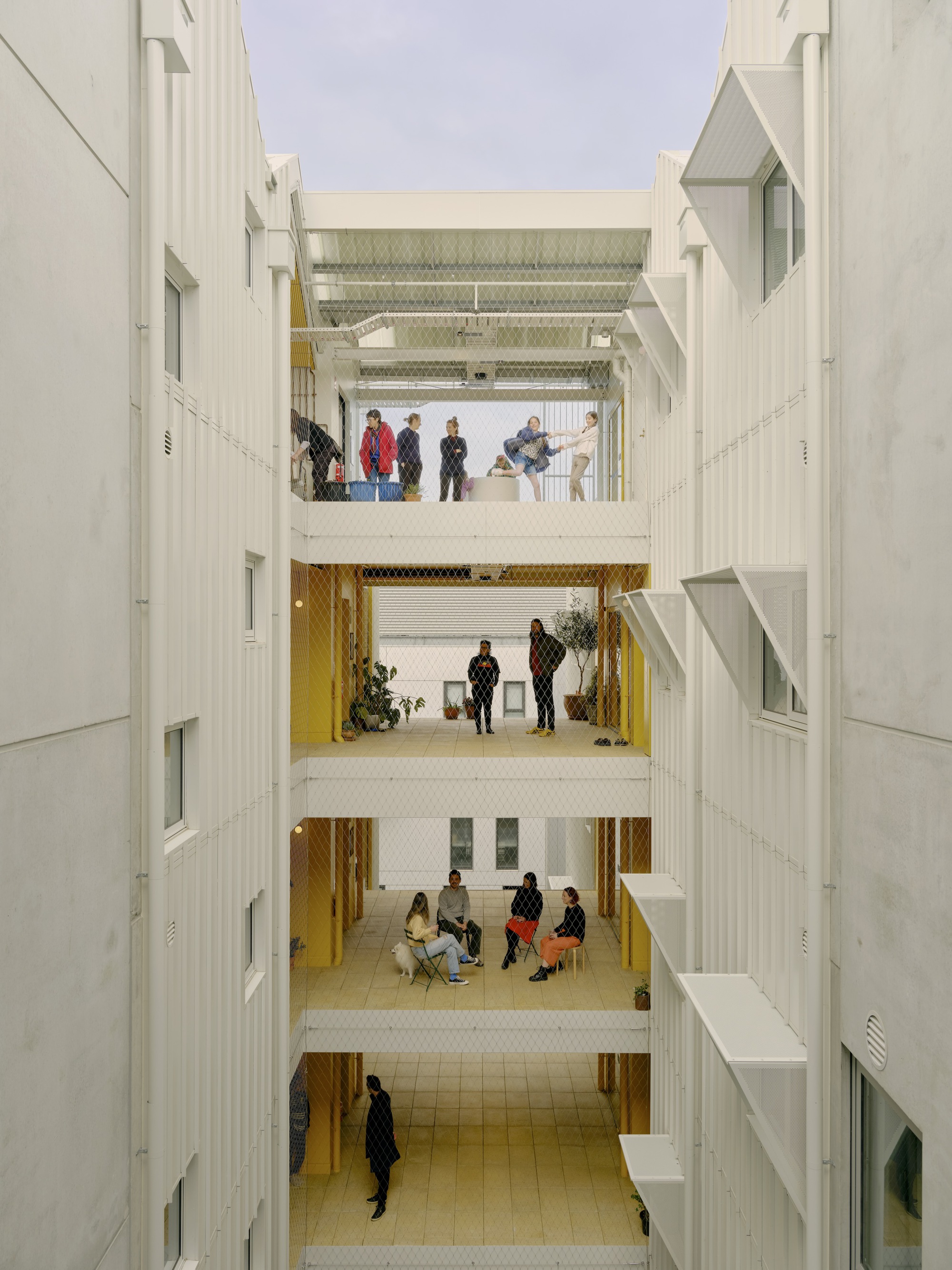 Austin Maynard Architects 澳洲集合住宅 Brunswick ParkLife 串聯各層的明亮中庭。建築師將傳統的走廊，擴展為寬敞的「空中街道」，足以擺放桌椅、植栽，促成不期而遇的交流。通透的網狀安全護欄（隱形鐵窗），取代了冰冷的實牆隔閡，讓光線與空氣自由流動，也讓鄰里間的視線與互動毫無阻礙