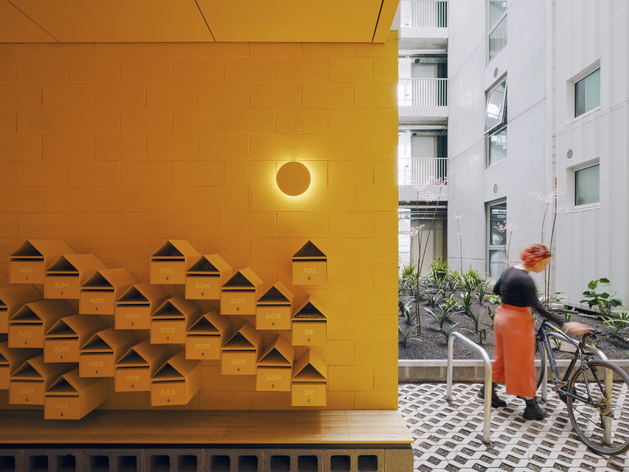 Austin Maynard Architects 澳洲集合住宅 Brunswick ParkLife 設計細節在入口處便清晰可見，暖黃色的塗砌磚牆成為視覺焦點，牆上排列著數十個訂製的金屬信箱，可愛的房屋輪廓，與建築頂層的鋸齒狀天際線相互呼應。地面採用的透水性鋪面，以及一旁的自行車停放架，皆是此處倡導永續生活方式的低調宣言