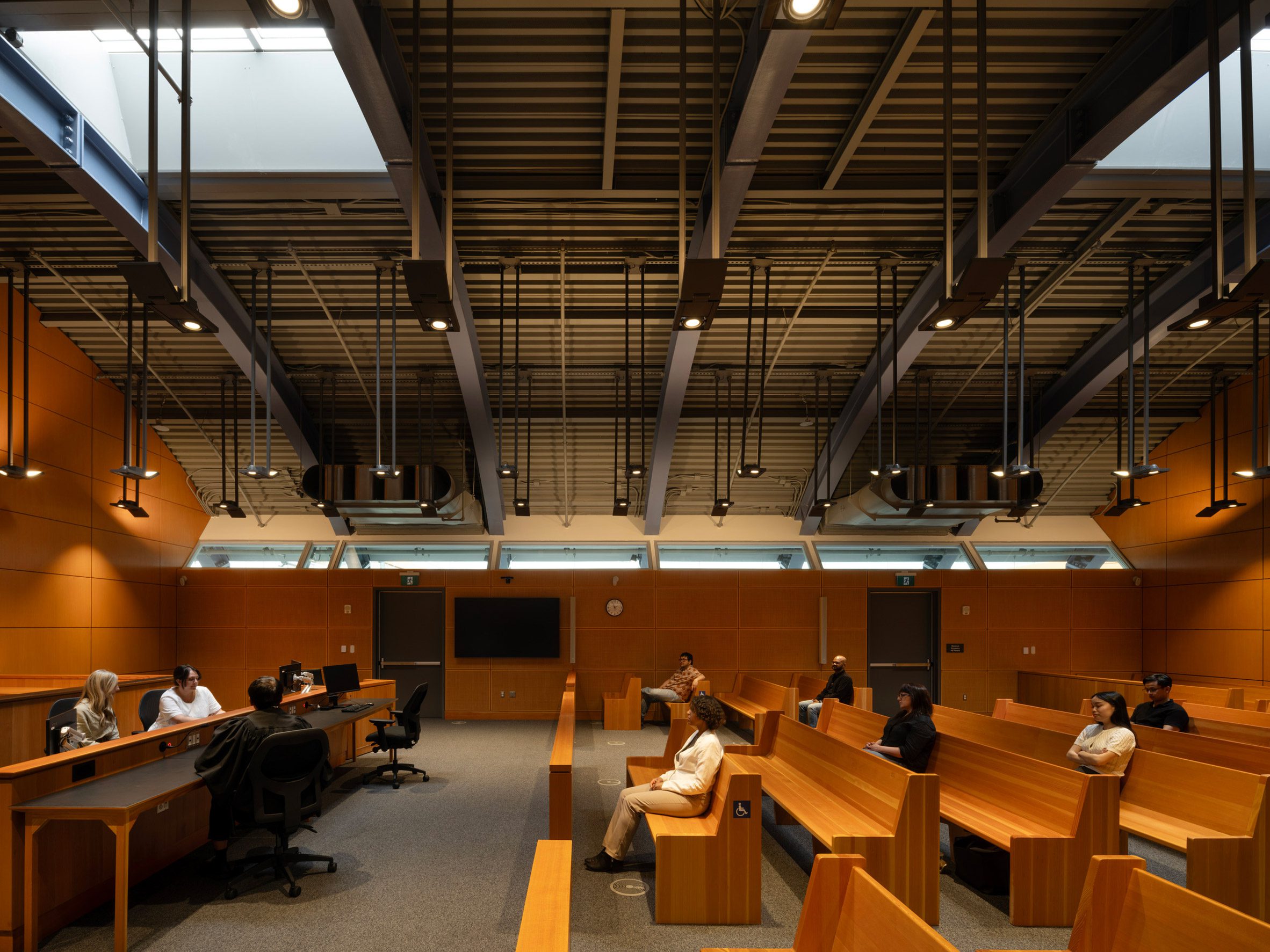 Richard Rogers RSHP 加拿大 St Lawrence Market North Toronto 在拱頂天窗的自然採光下，法庭空間呈現一種靜謐氛圍。溫潤的木質傢俱、辦公椅與牆面，和天花板暴露的深色金屬結構，在材質上構成冷暖對話。從法官席到旁聽席的序列安排，清晰地界定出此一市民空間所應用的莊重機能與秩序