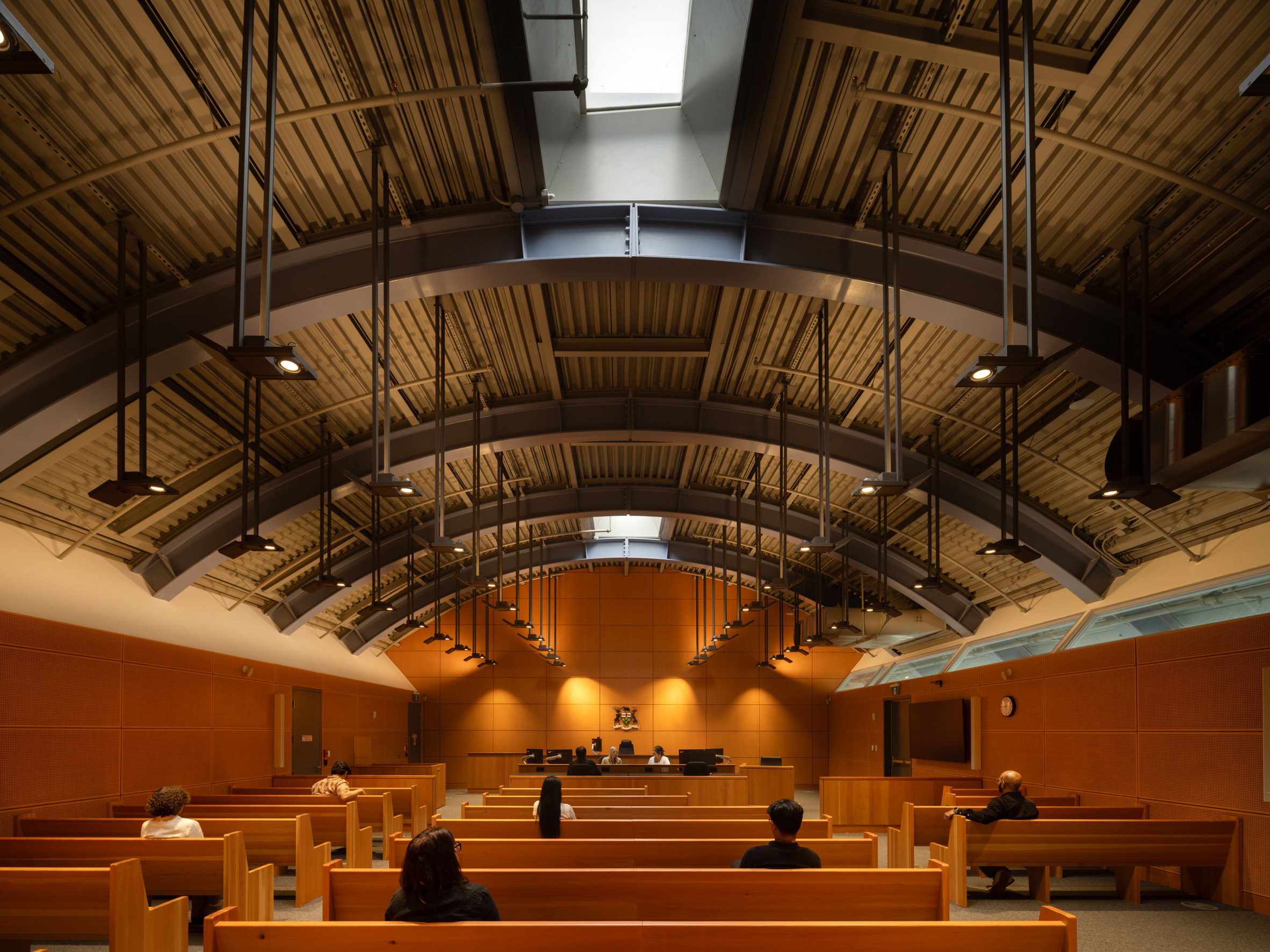 Richard Rogers RSHP 加拿大 St Lawrence Market North Toronto 位於建築上層的市級法庭，其空間結構直接對應著建築外觀的宏偉拱頂。天光自頂部天窗悄然灑落，映照在序列整齊的木質旁聽席長椅，以及鋪滿牆面的吸音板上。外露的金屬穹頂與懸吊燈具，為莊嚴的司法場域帶來一絲現代工業氣息